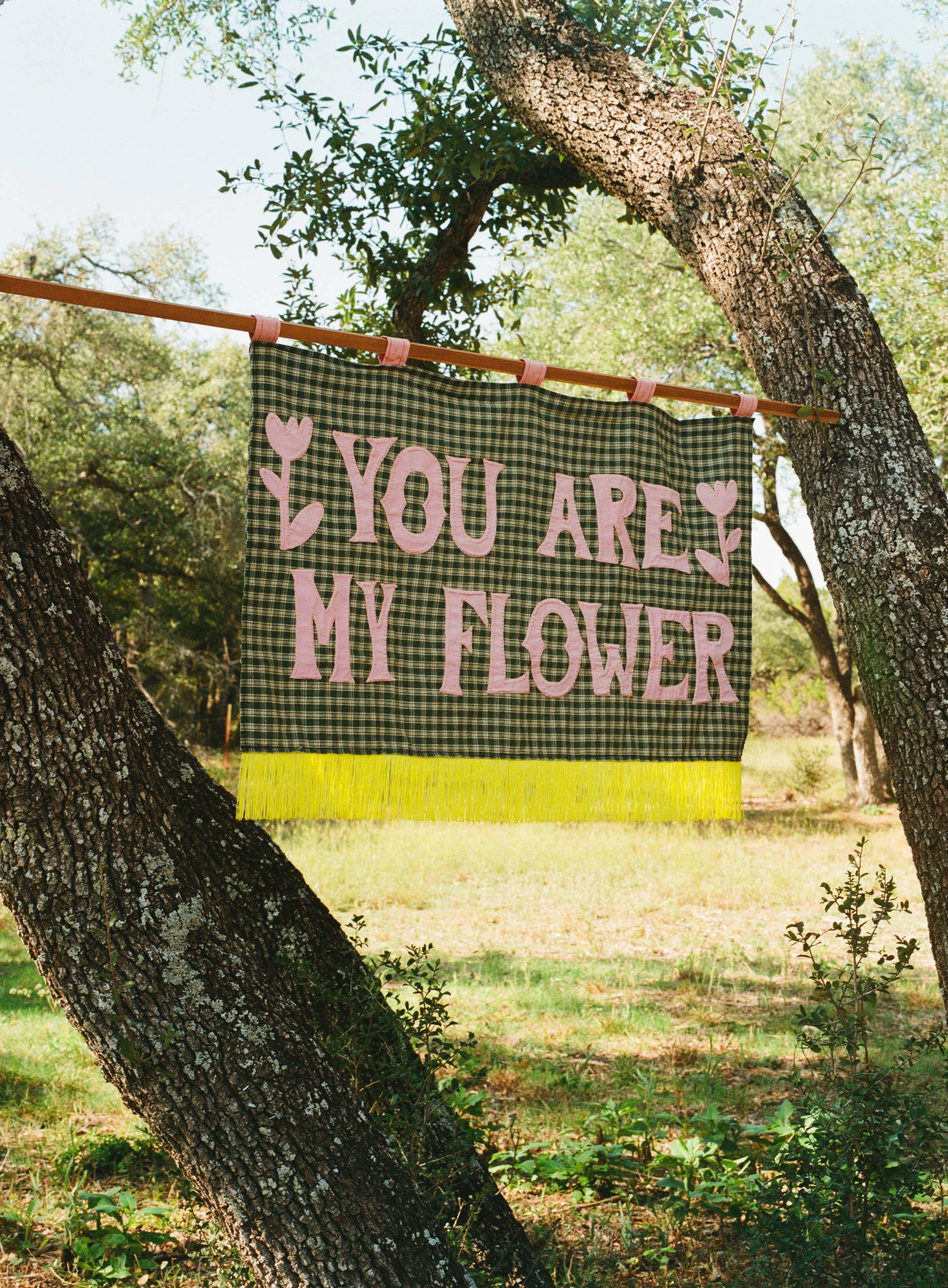 A decorative fabric sign hanging on a wooden pole tied to tree branches outdoors. The sign has a green and black checkered background with pink felt letters and hearts, reading 'YOU ARE MY FLOWER'.