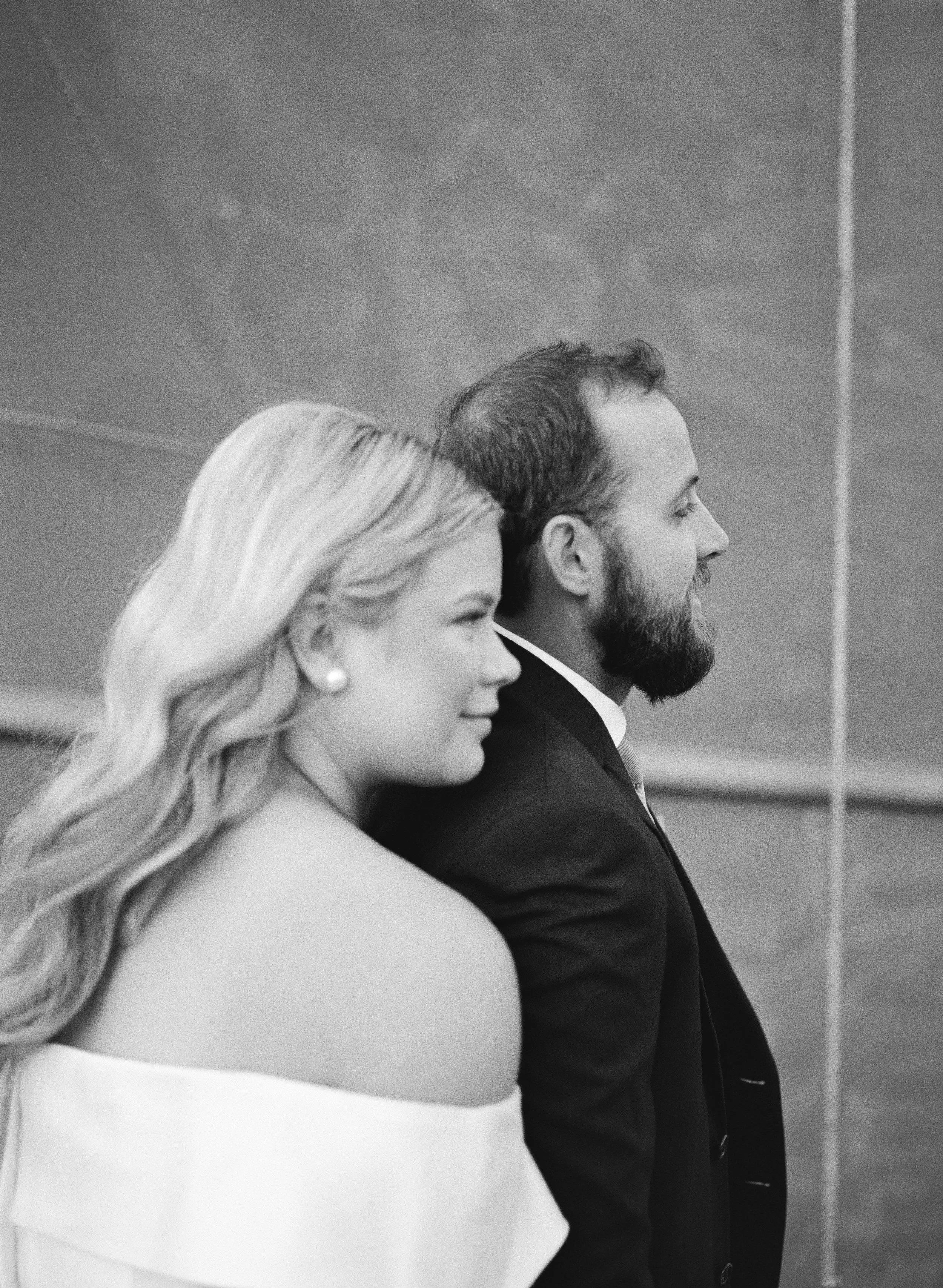 A black and white photo of a woman and a man standing side by side, looking forward. The woman has long, wavy hair and is wearing an off-the-shoulder dress and pearl earrings. The man has a beard and short hair, wearing a suit and tie, with a wooden 