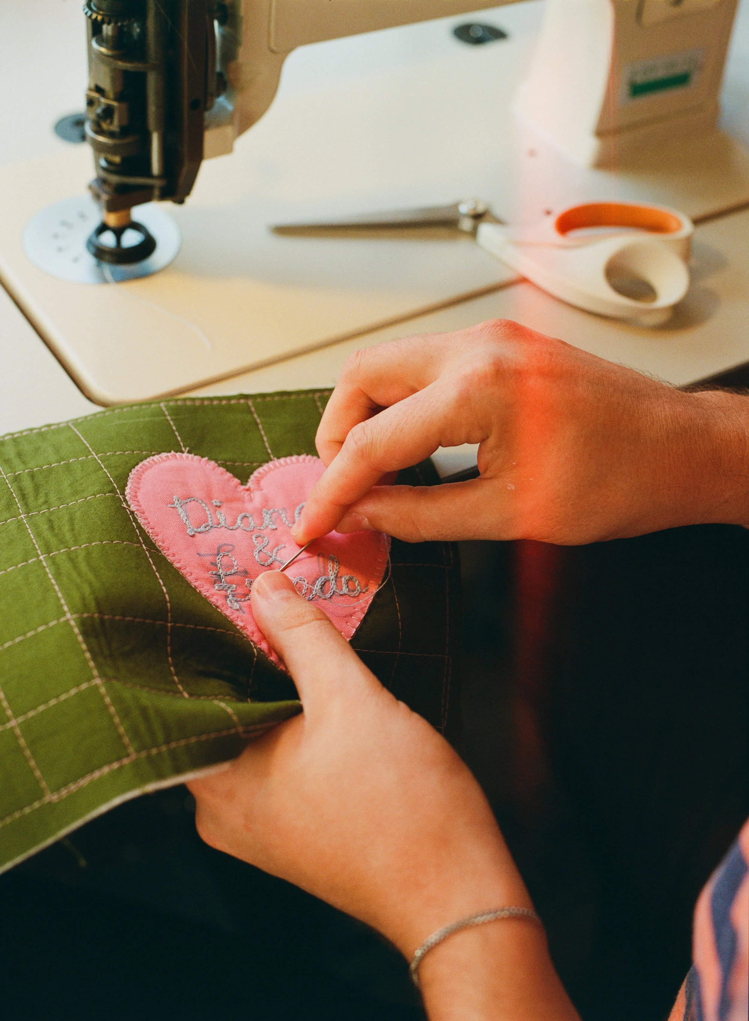 Person hand sewing an embroidered pink heart patch onto green fabric using a sewing needle, with sewing machine, scissors, and thread near them on the table.