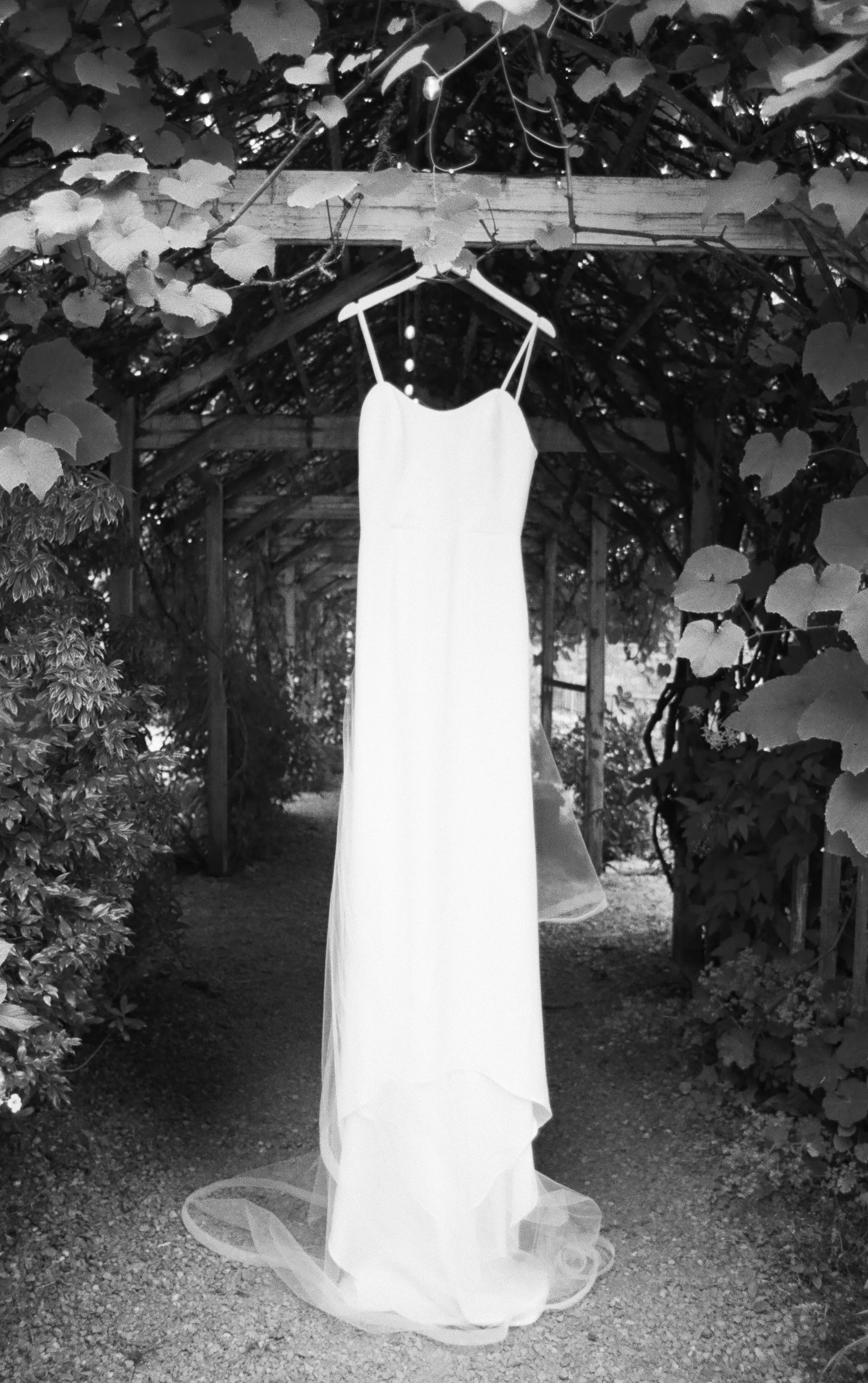 A white wedding dress hanging on a coat hanger from a wooden beam in a garden pathway.