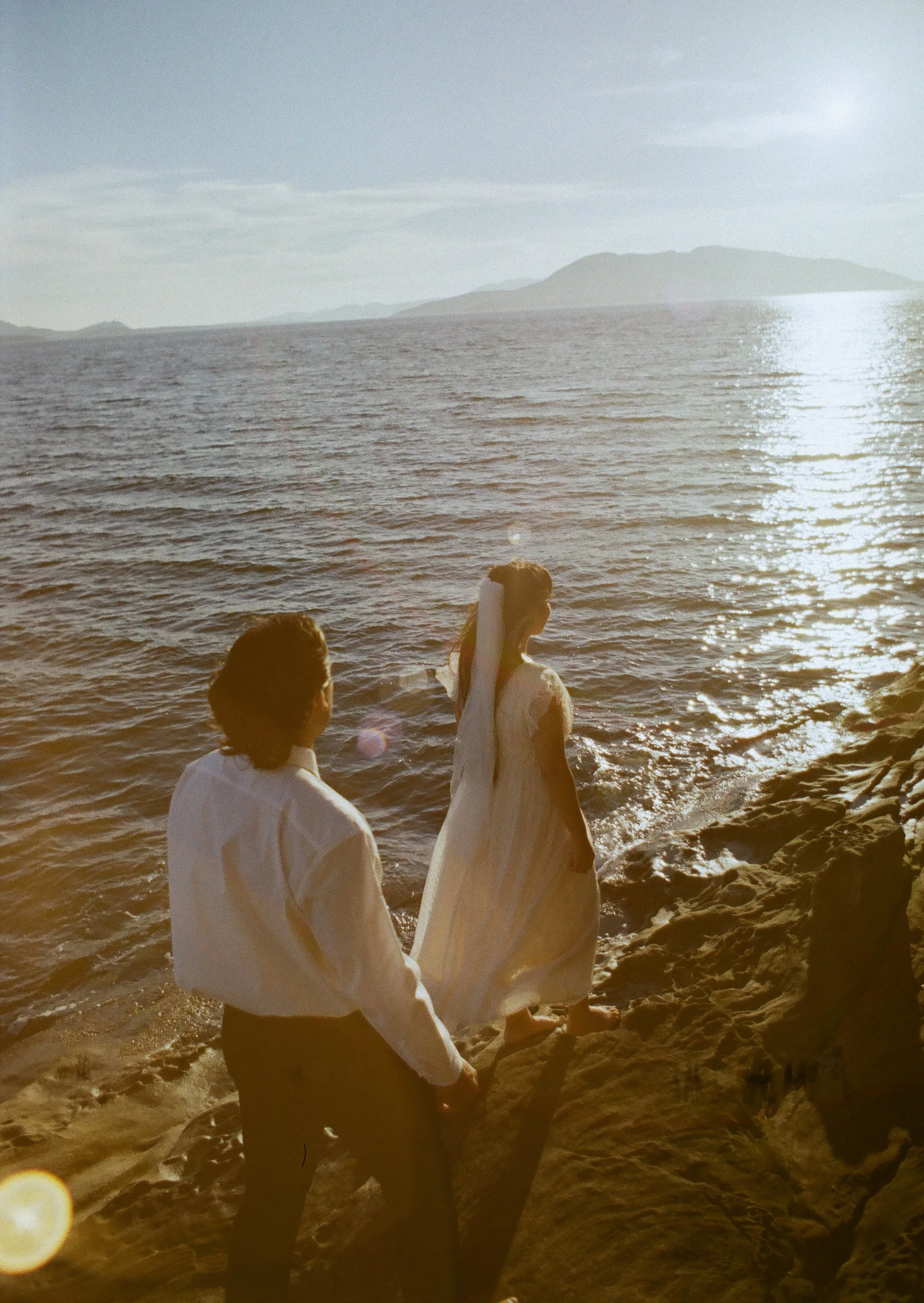 A woman and a man standing on rocks by the ocean during sunset. The woman wears a white dress, and the man wears a white shirt and dark pants. The woman faces the ocean, holding her hair, while the man looks at her.