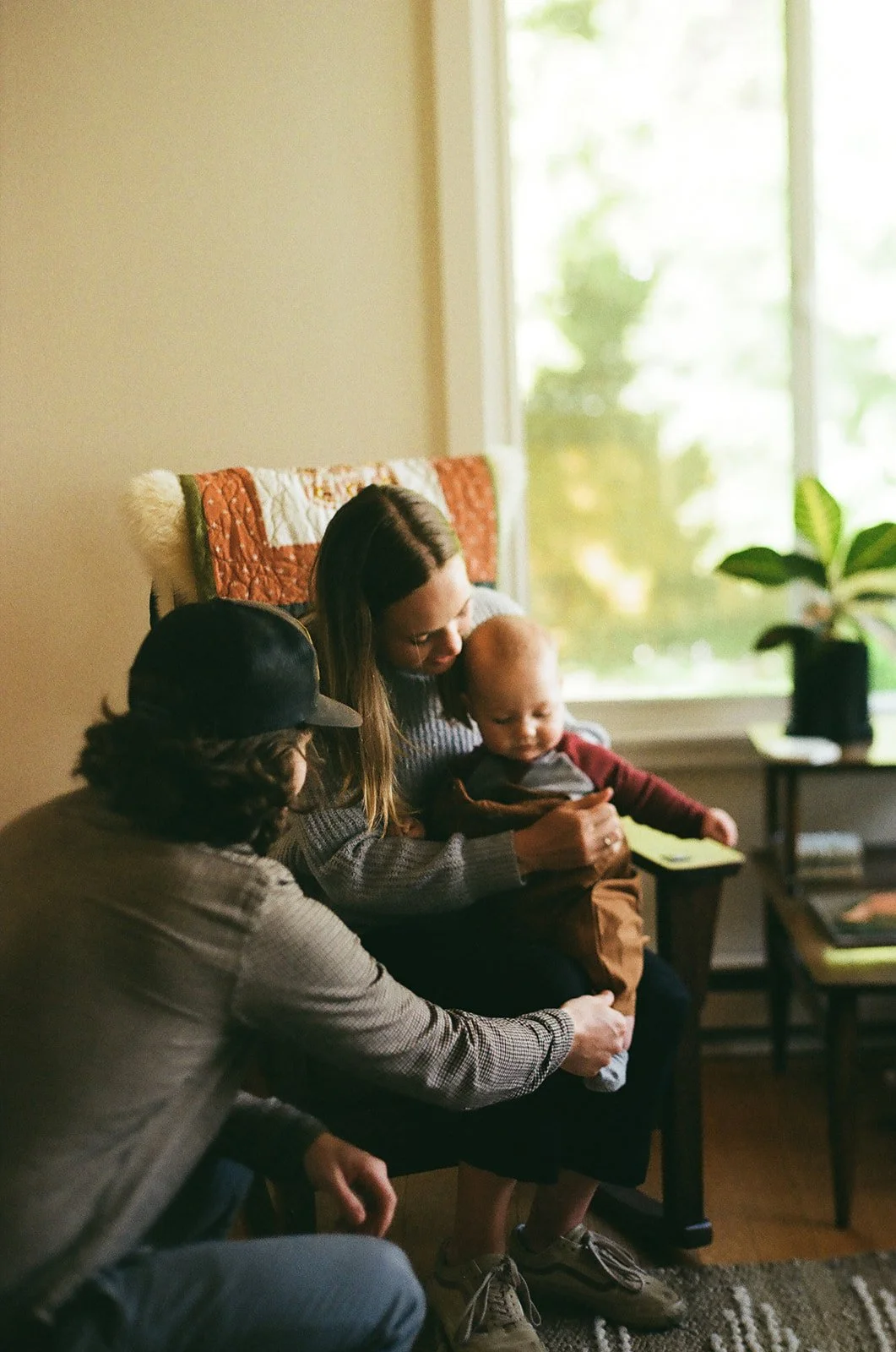 Woman sitting in a chair with a young child on her lap while another person kneels nearby, all focused on the child, inside a cozy room with a window and houseplant.