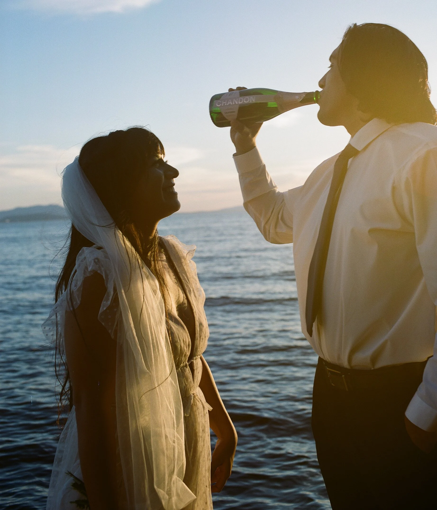 A man and woman by the water at sunset, the man is drinking from a bottle, the woman is smiling.
