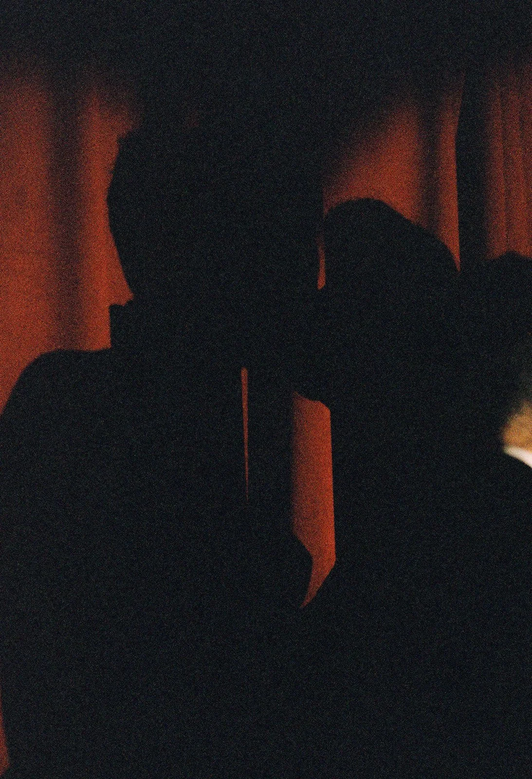 A dark, shadowy silhouette of a person taking a selfie in front of a curtain, with a faint red glow in the background.