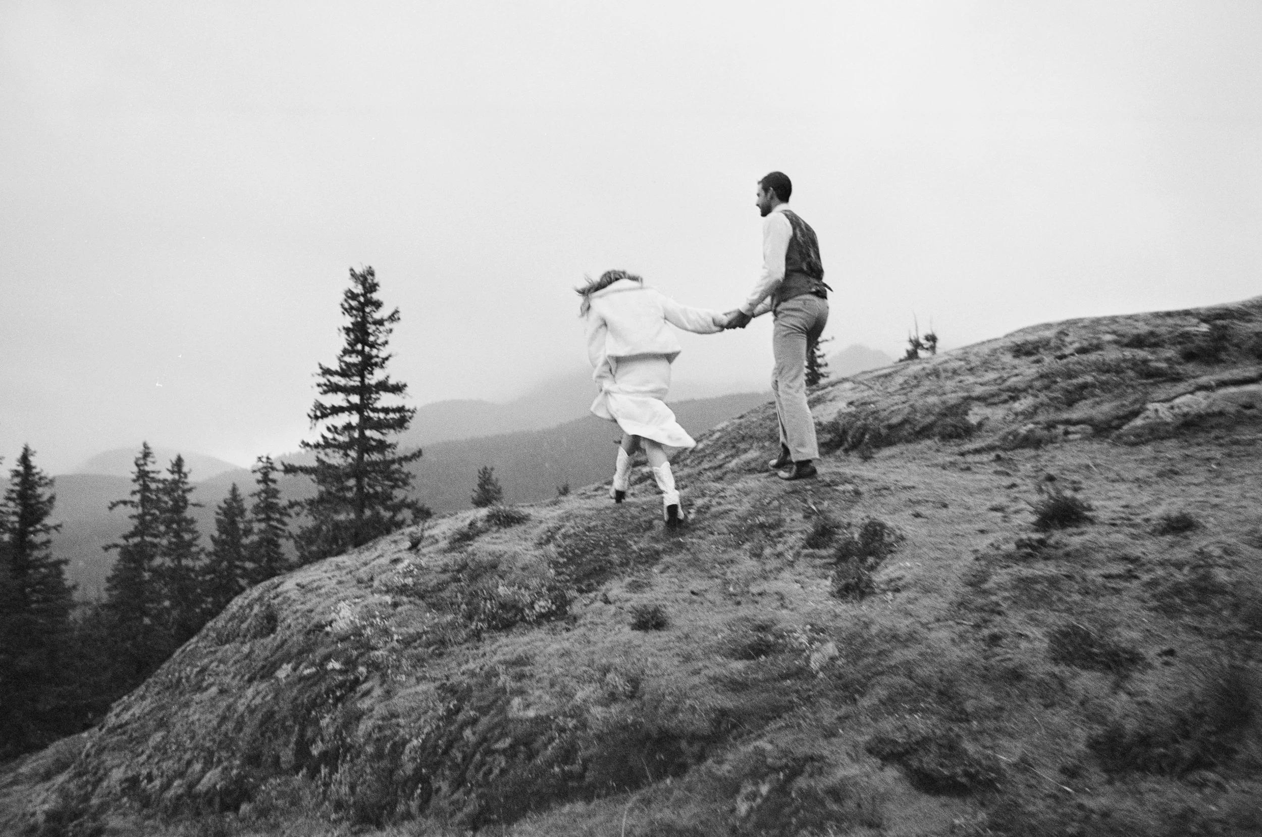 A man and a girl holding hands on a rocky hillside with a forest and mountains in the background, black and white photo.