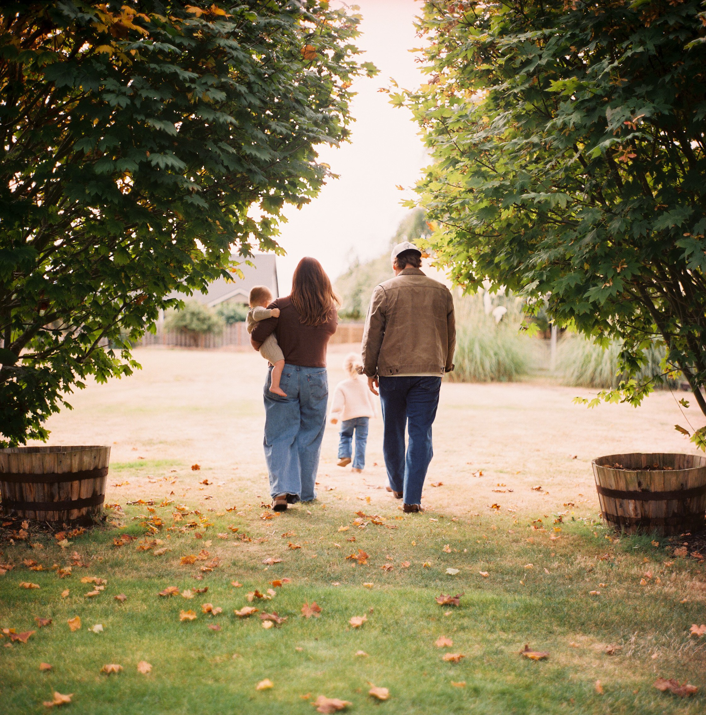 Family walking outdoors through a garden, viewed from behind, with large leafy trees and a grassy area leading to a more open space in the background.