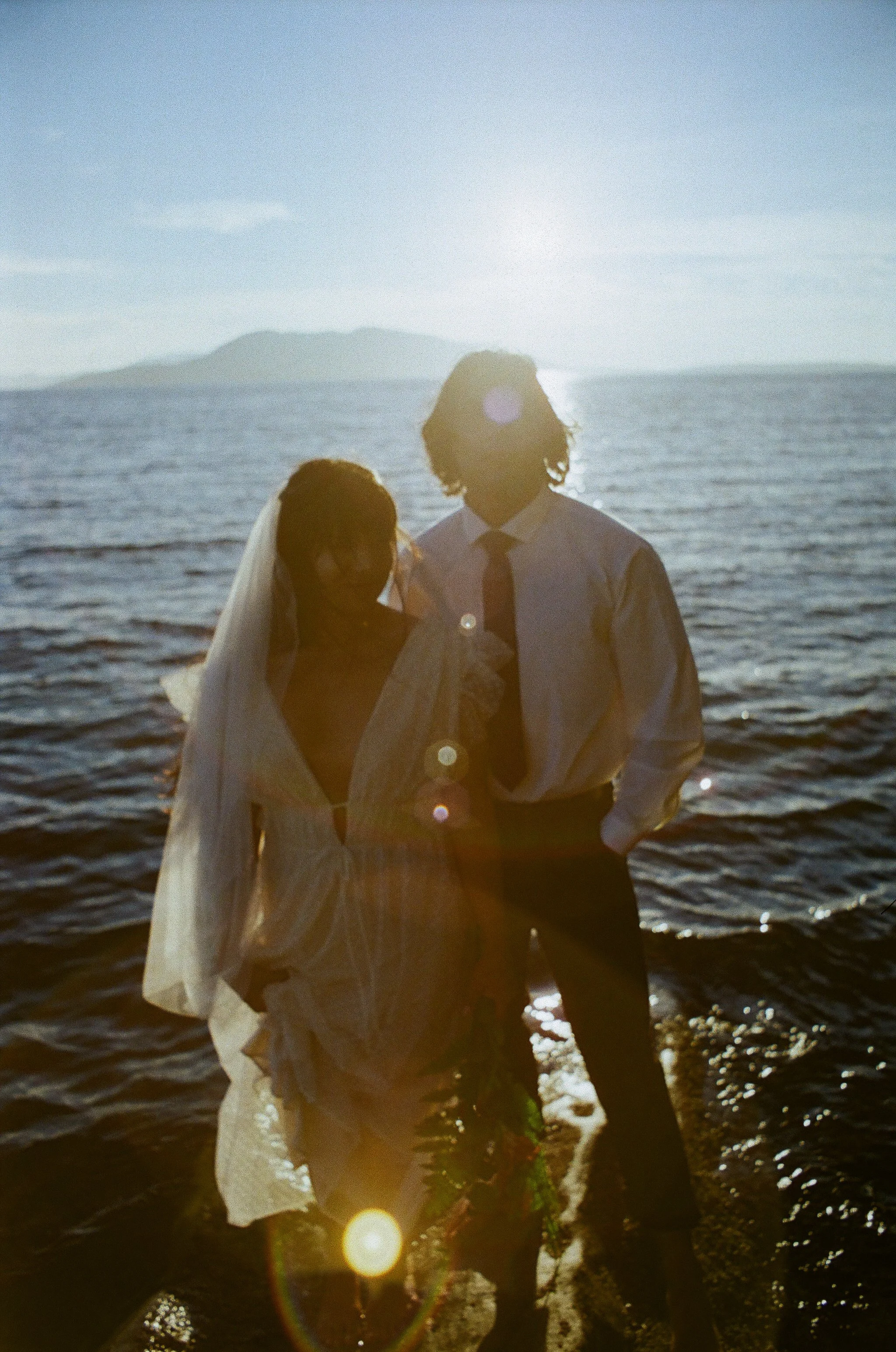A couple dressed in wedding attire, holding a bouquet, standing in water with the sun setting behind them near a body of water and an island in the background.