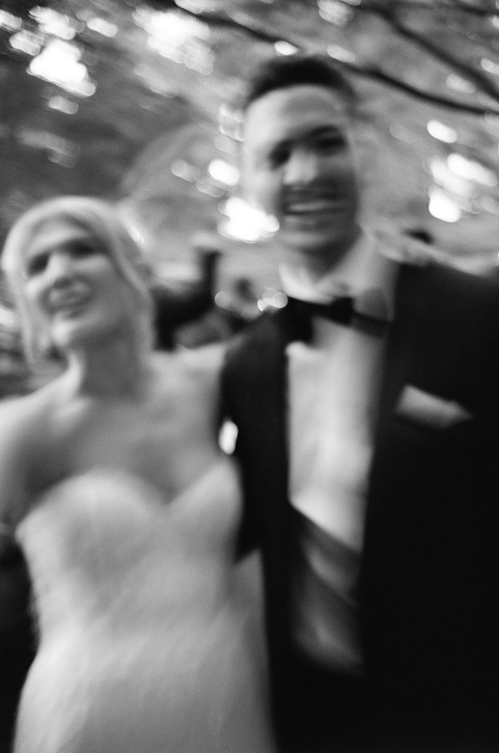 A blurry black and white photo of a smiling woman in a wedding dress and a man in a tuxedo, likely at their wedding reception.