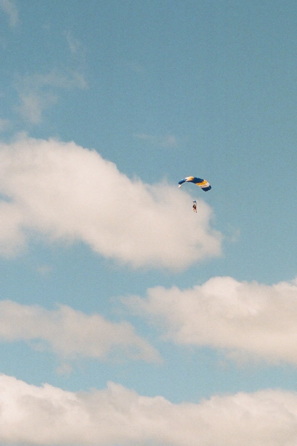 A person skydiving with a yellow, blue, and orange parachute against a blue sky with white clouds.