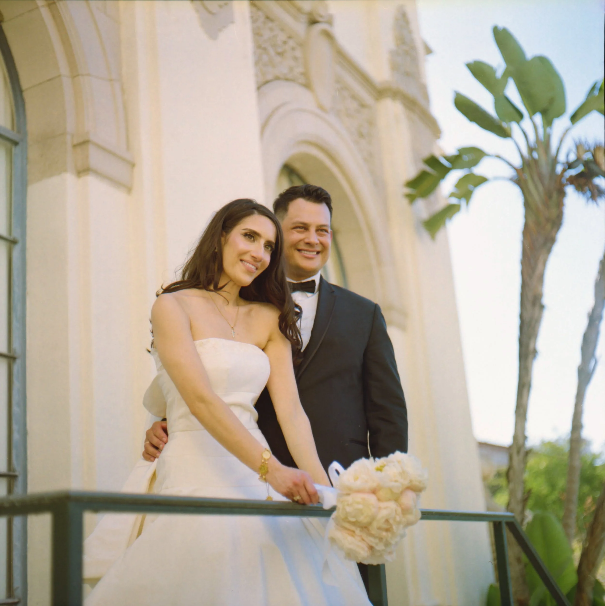 A bride and groom standing on a balcony outside a building, smiling. The bride is holding a bouquet of flowers and wearing a strapless wedding gown, and the groom is in a tuxedo with a bow tie. A palm tree is visible in the background.