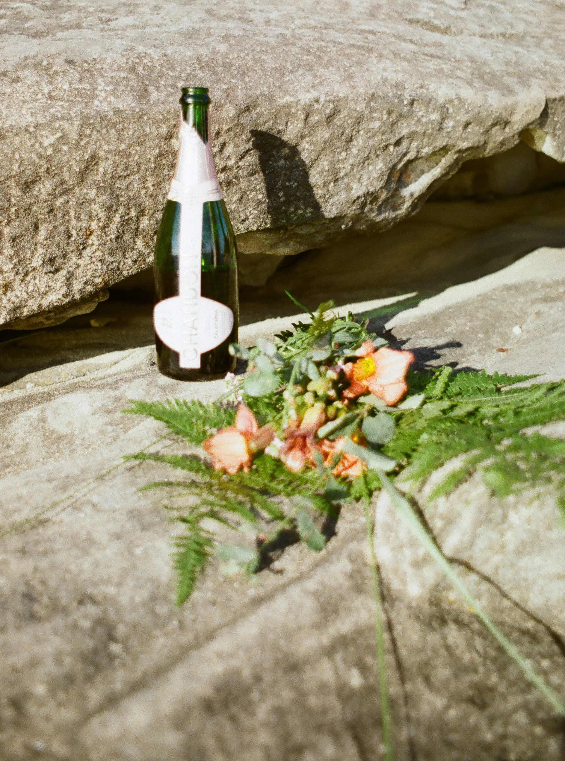 A green glass champagne bottle and a small bouquet of flowers and greenery placed on a stone surface outdoors.