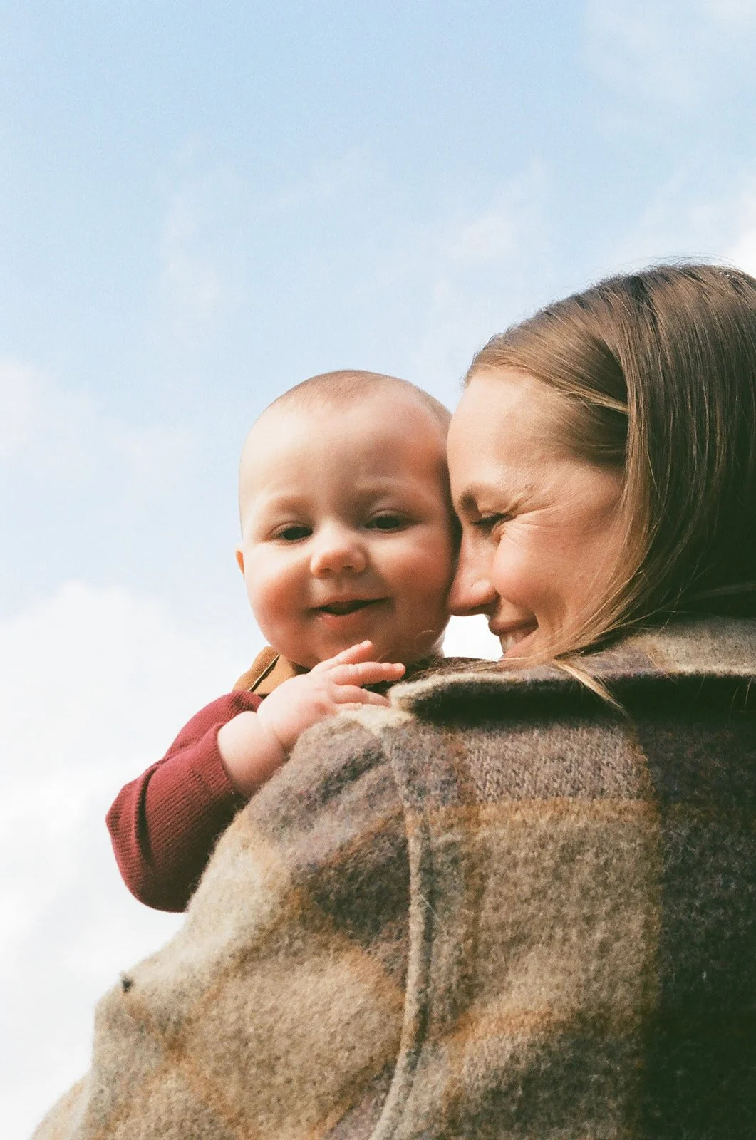 A woman holding a baby close, both smiling and making contact with their faces against a blue sky with a few clouds.