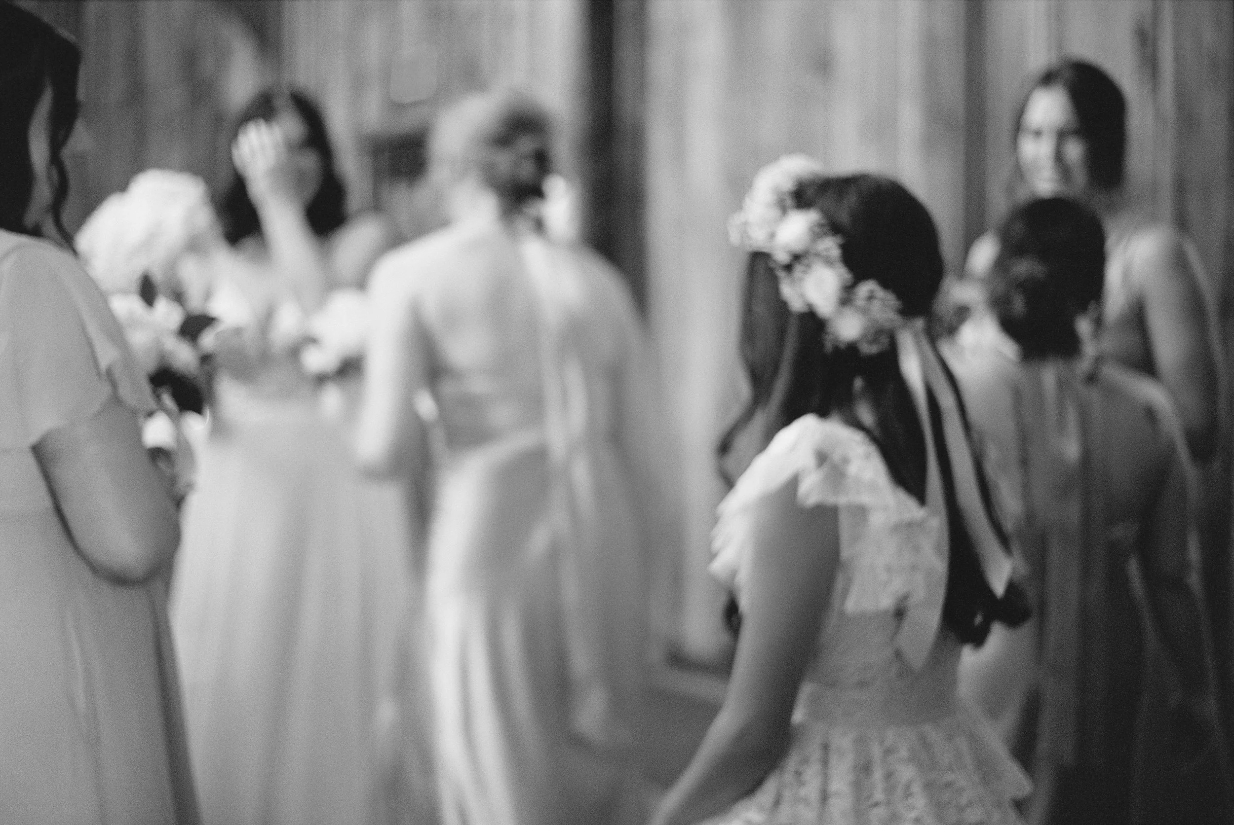 A wedding reception scene in black and white, showing a bride with a floral crown and lace dress standing among guests in a rustic setting.