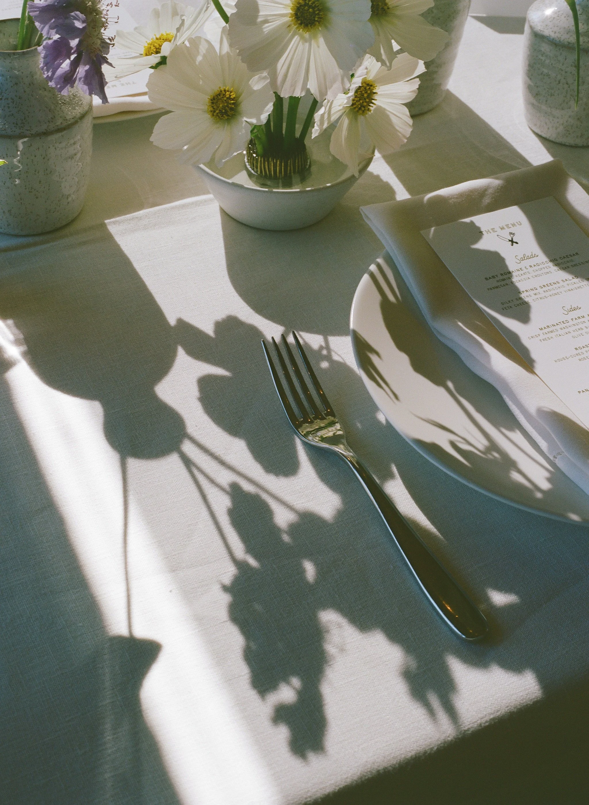 A table setting with white flowers in vases, a white plate with a menu, a fork, and decorative shadows cast by the flowers and tableware.