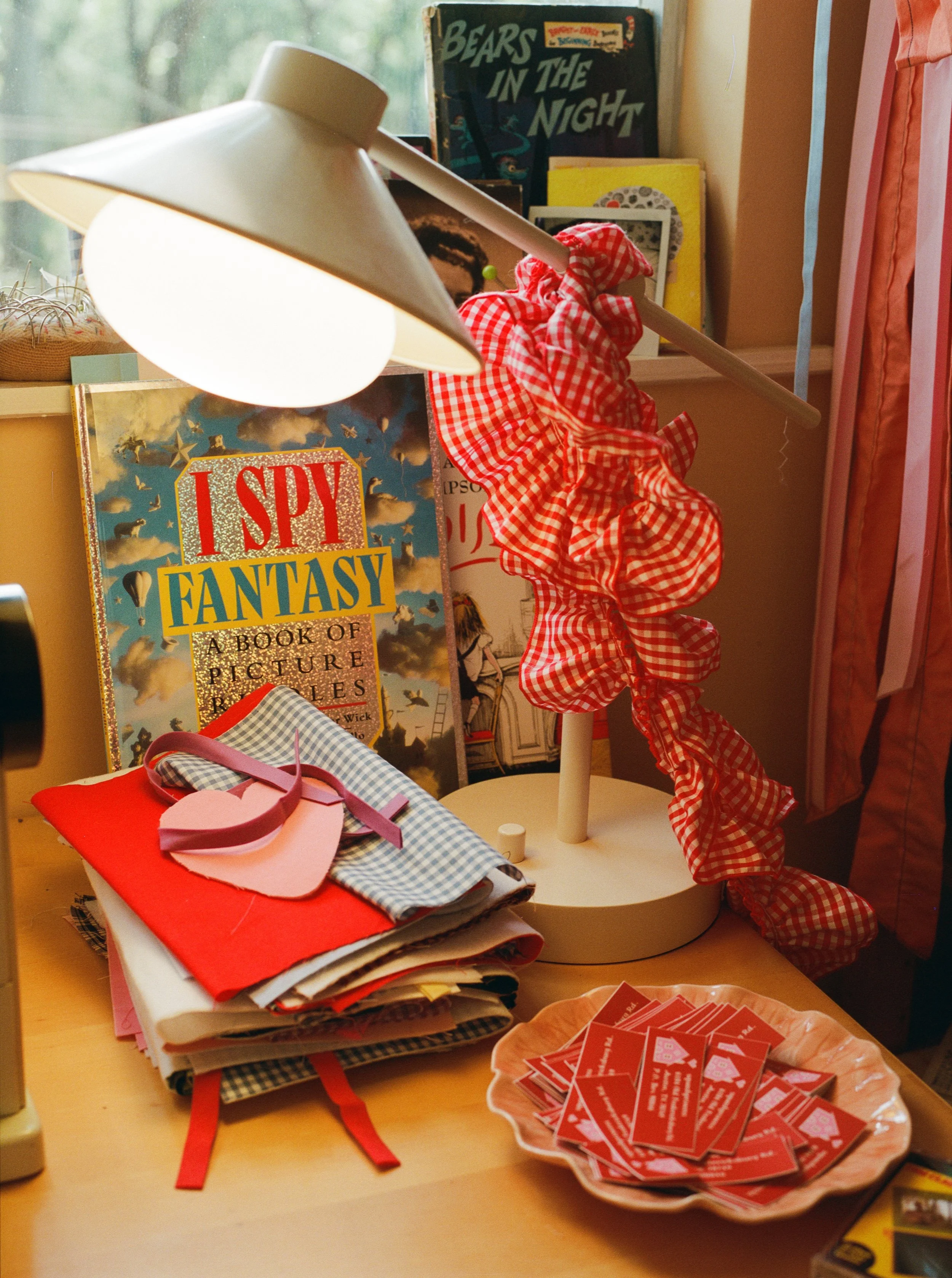 A cluttered desk with a stack of fabric and paper materials, a paper heart with a ribbon, a bowl filled with red gift tags, a book titled "Spy Fantasy," a decorative lamp, and a dress form with a red and white checkered ruffle, located next to a wind