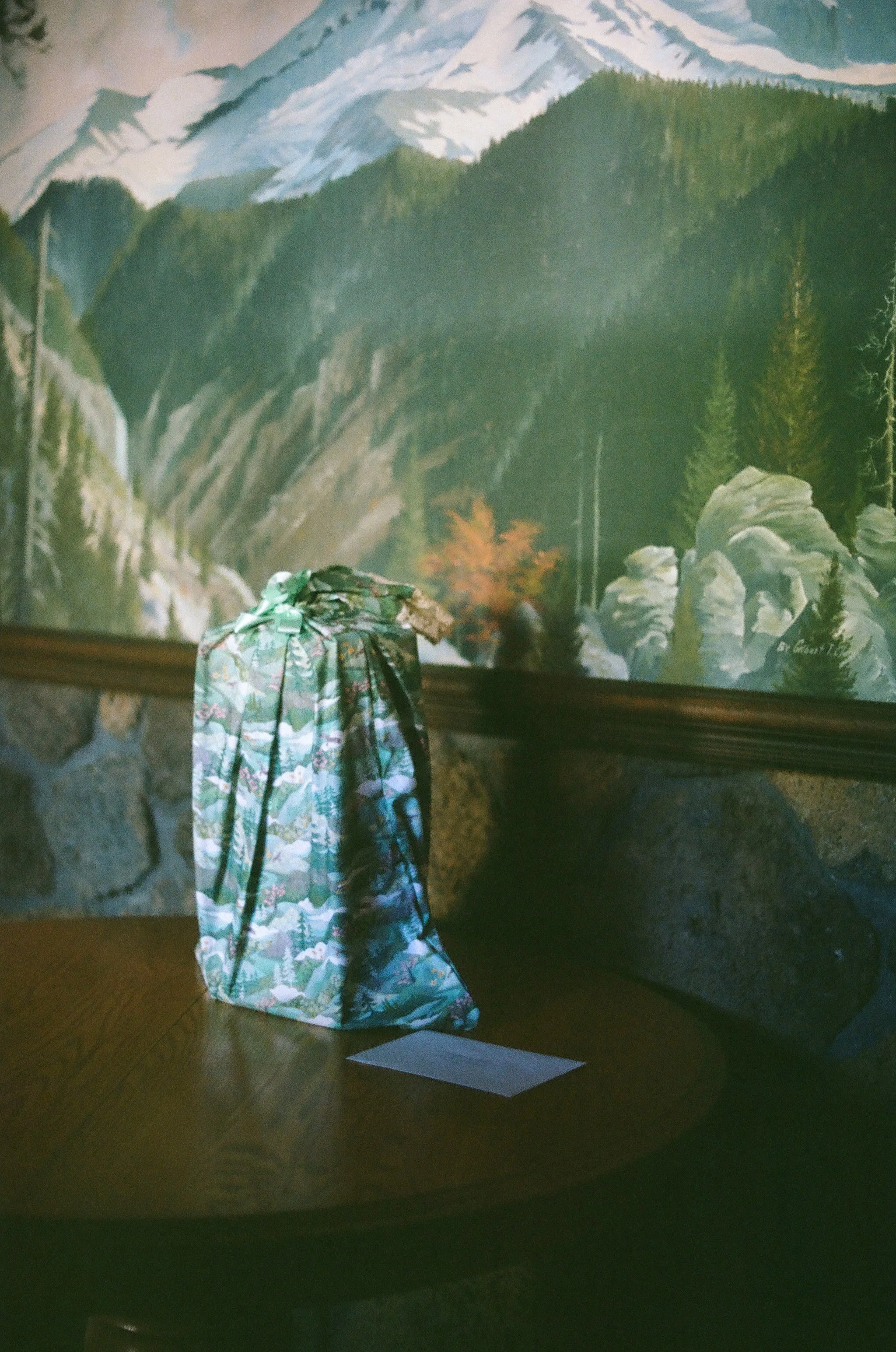 A gift wrapped in floral paper with a ribbon, placed on a wooden table in front of a mountain landscape painting.