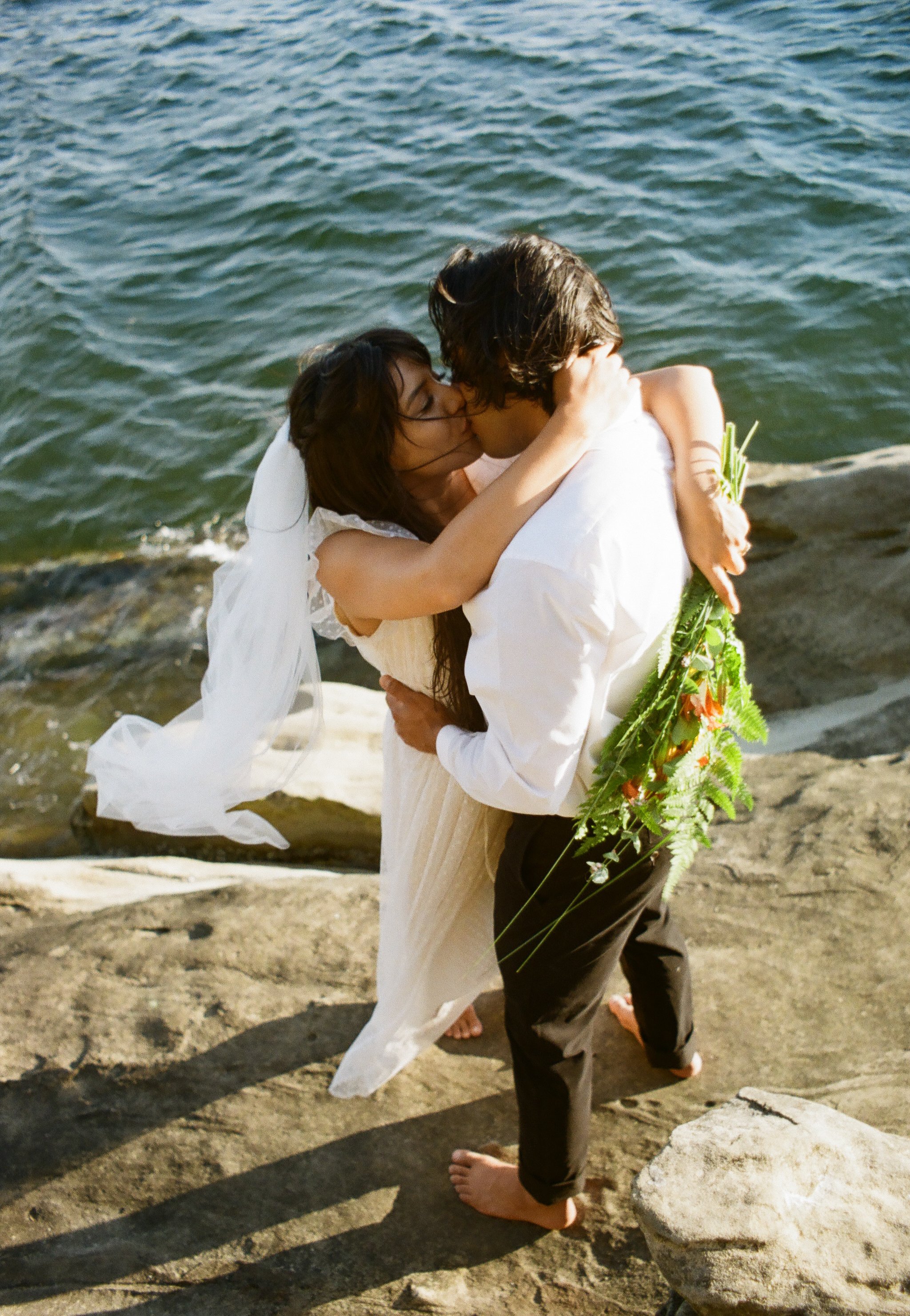 A couple in wedding attire embracing and kissing on a rocky shoreline near the water, with the woman holding a bouquet of flowers.