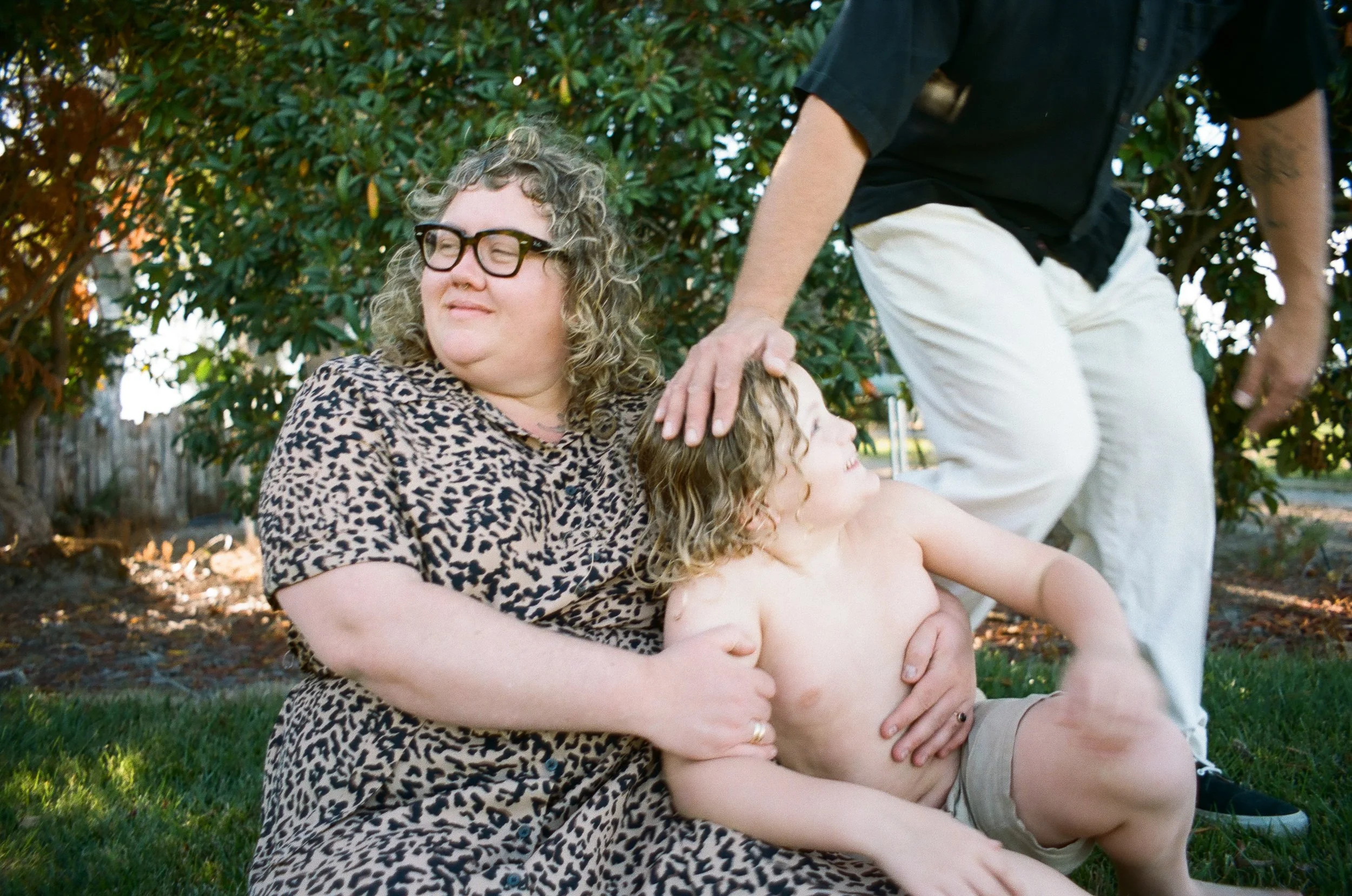 A woman with curly blonde hair, glasses, and a leopard print dress is sitting on the grass, holding a shirtless young boy with curly blonde hair. A person in a black shirt and white pants is standing nearby, resting their hand on the boy's head. They