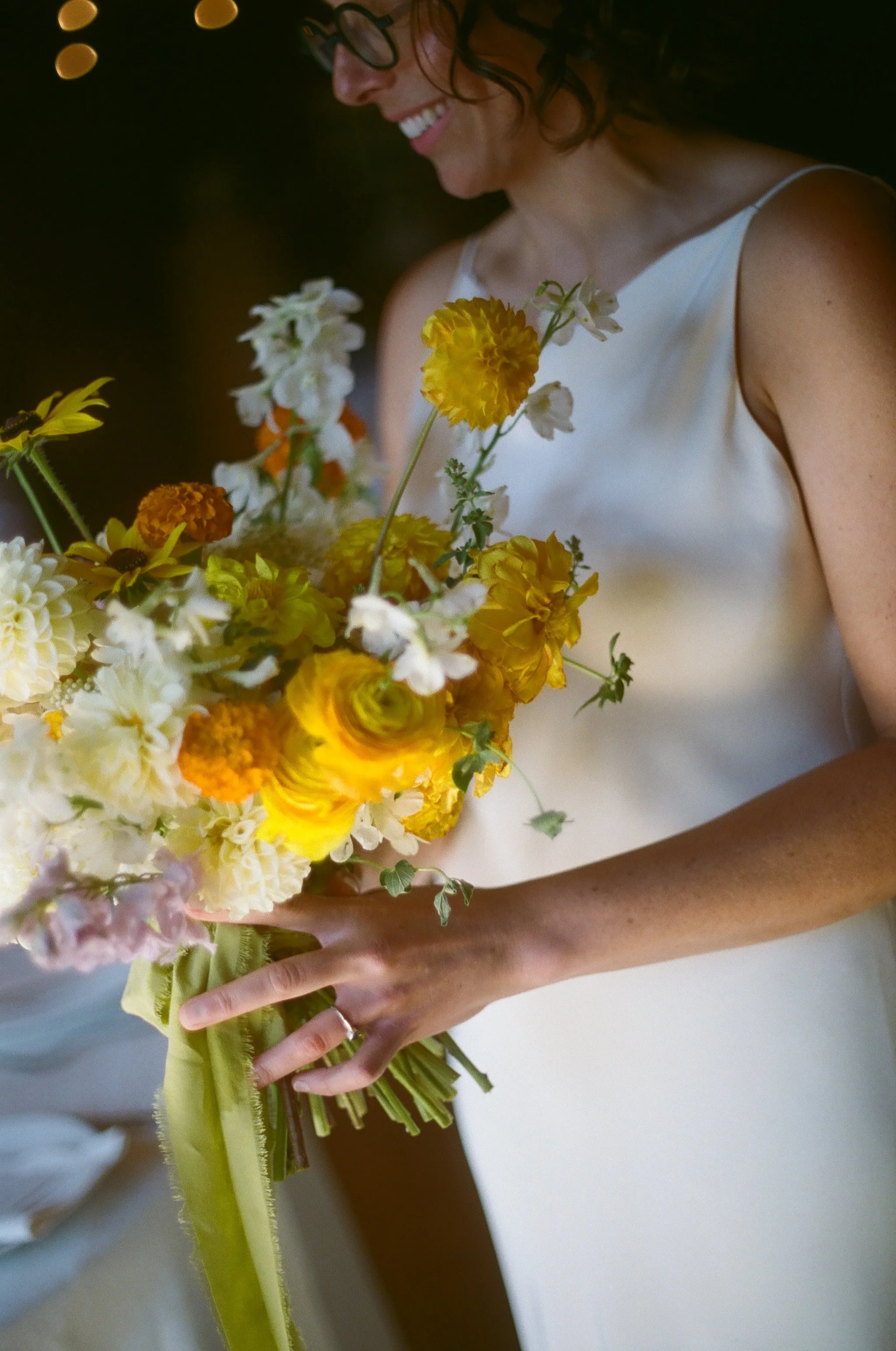 A woman in a white dress holding a large bouquet of yellow and white flowers, smiling and looking down.