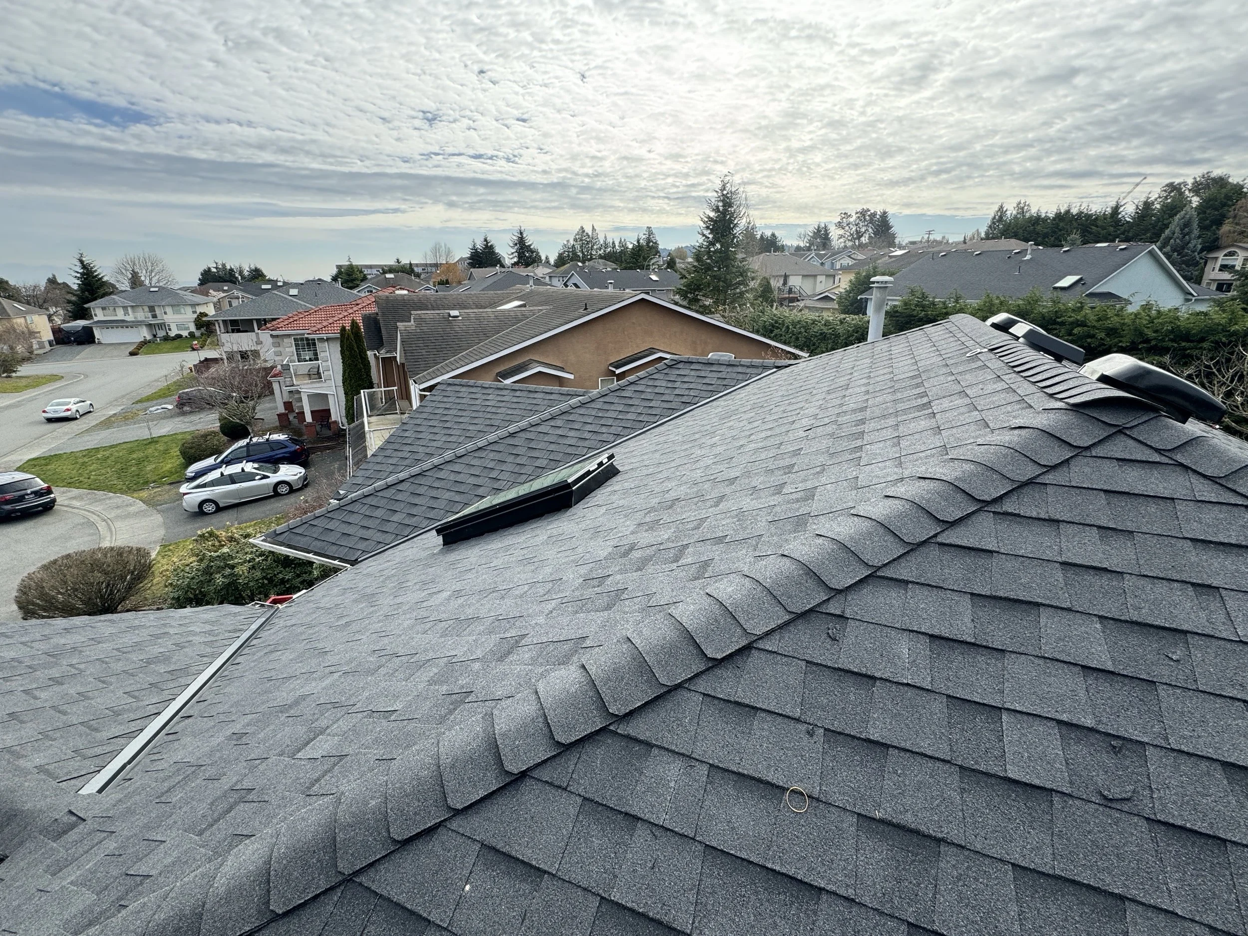 View of a neighborhood with houses with various roof styles and colors, with some cars parked on the street and in driveways, under a sky with scattered clouds.