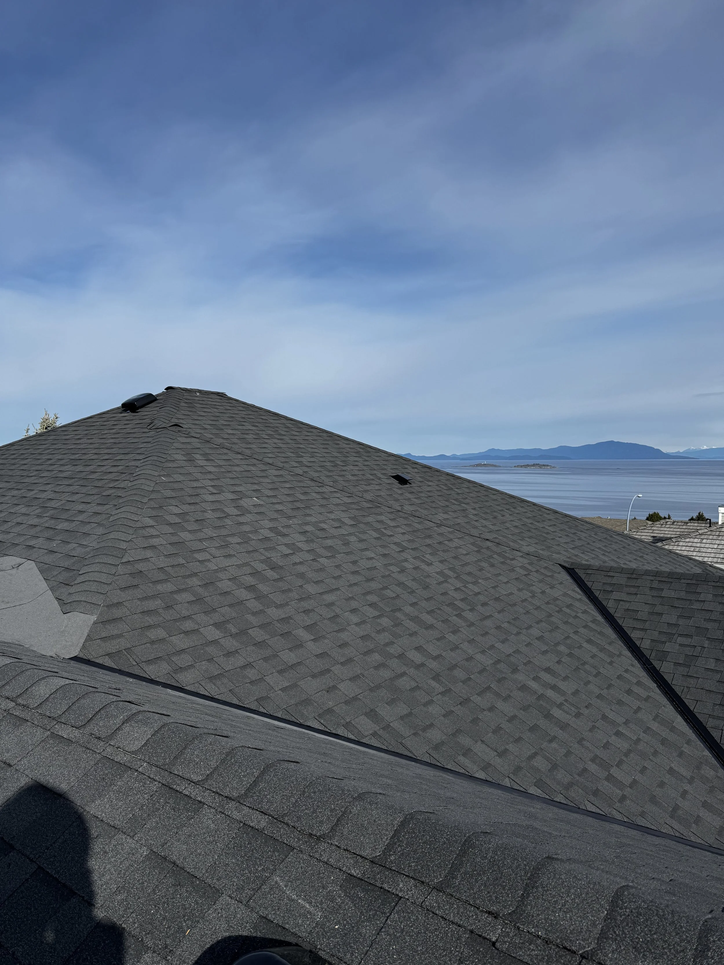 A rooftop with gray shingles, a chimney pipe, and vents, overlooking a body of water and mountains in the distance under a partly cloudy sky.