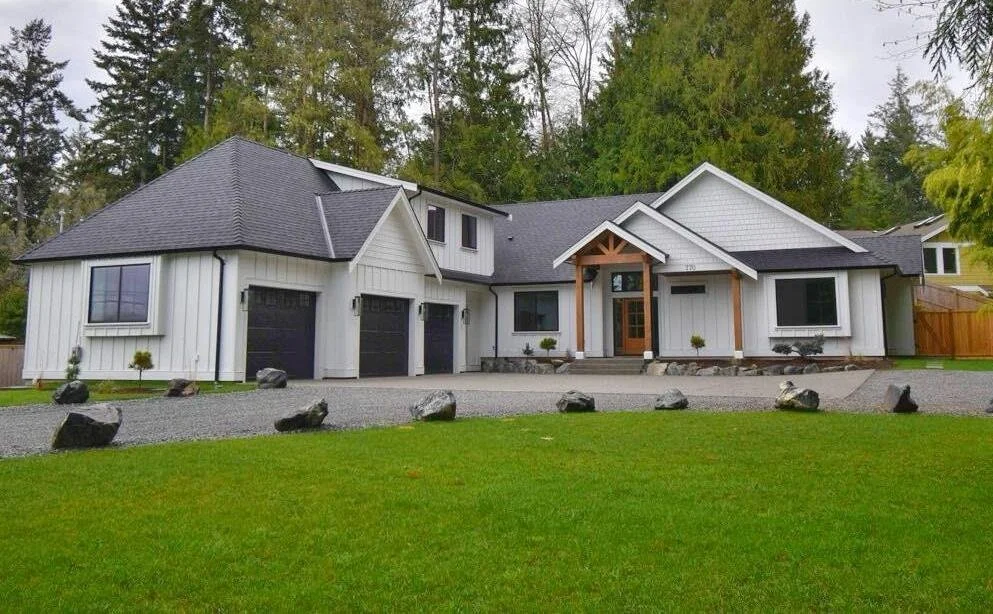 Modern white house with black garage doors, a black roof, and a front porch with wooden accents, surrounded by a green lawn, rocks, and trees in the background.