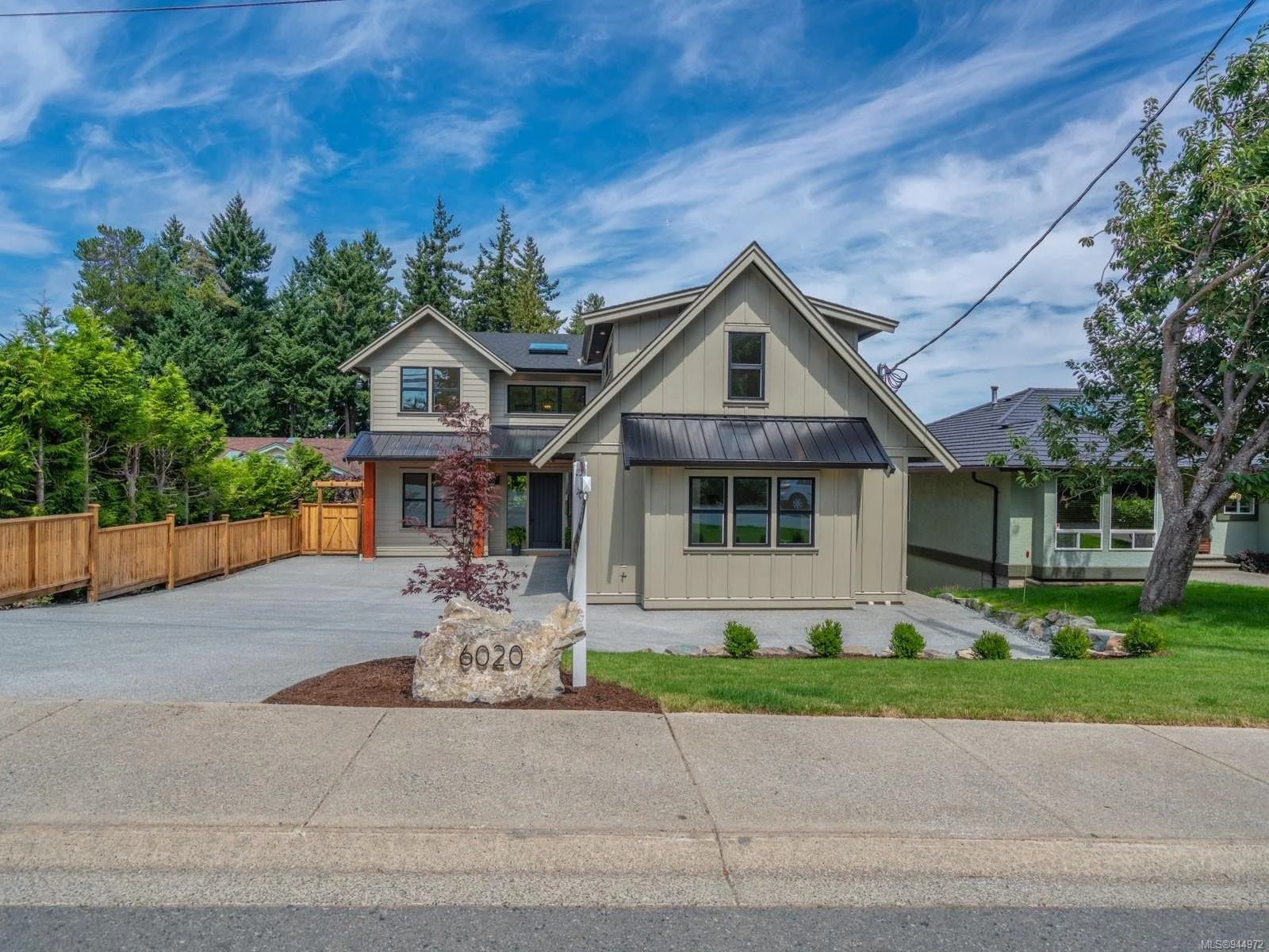 Modern two-story house with a gray exterior, black trim, and a black metal roof. It has a front yard with small bushes, a tree on the right, and a large rock with the house number 6020. The driveway is made of gravel, and there is a wooden fence on t