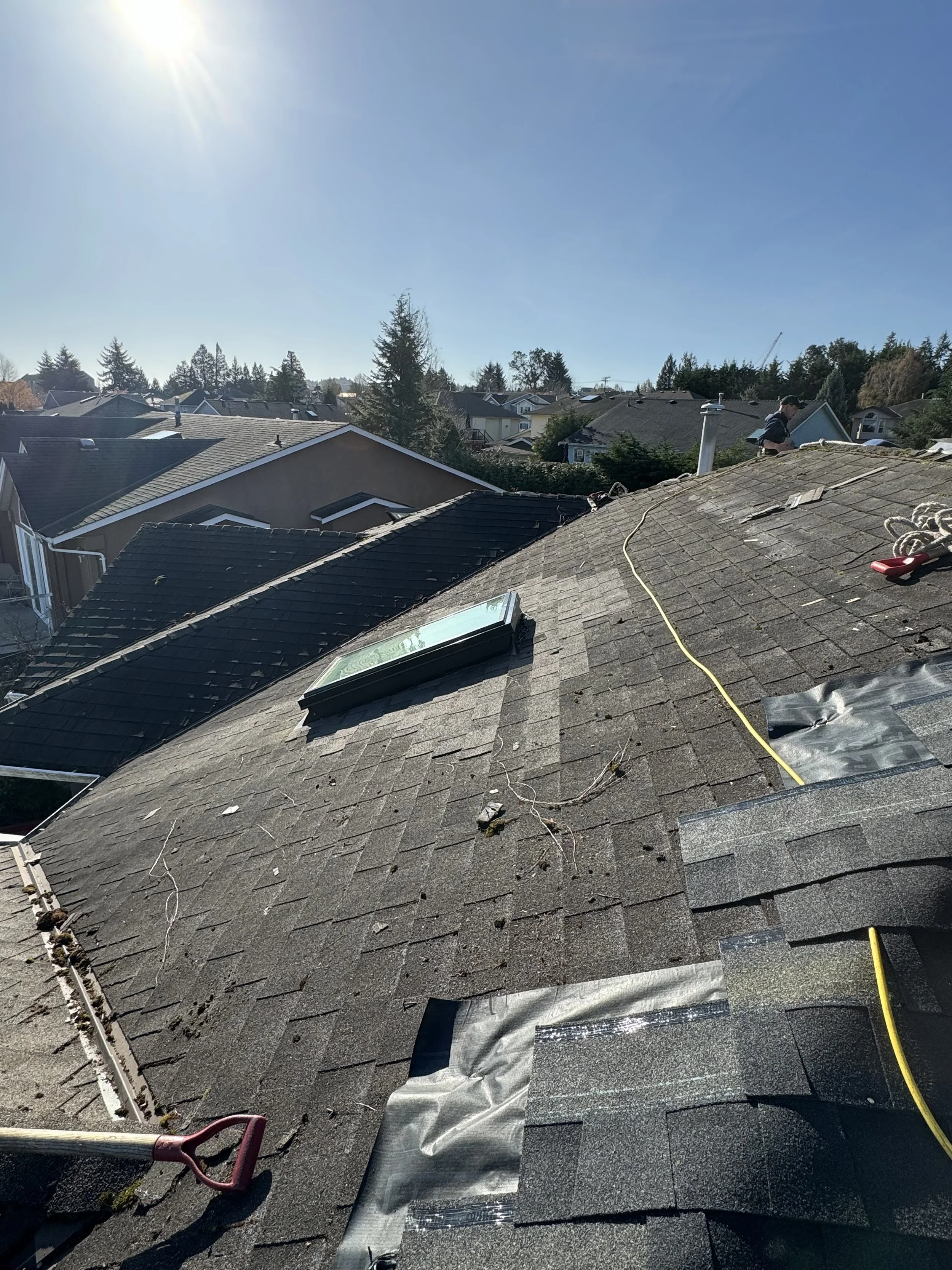 View of a rooftop with shingles, skylights, and a person working near the chimney under a clear sky with sunlight.