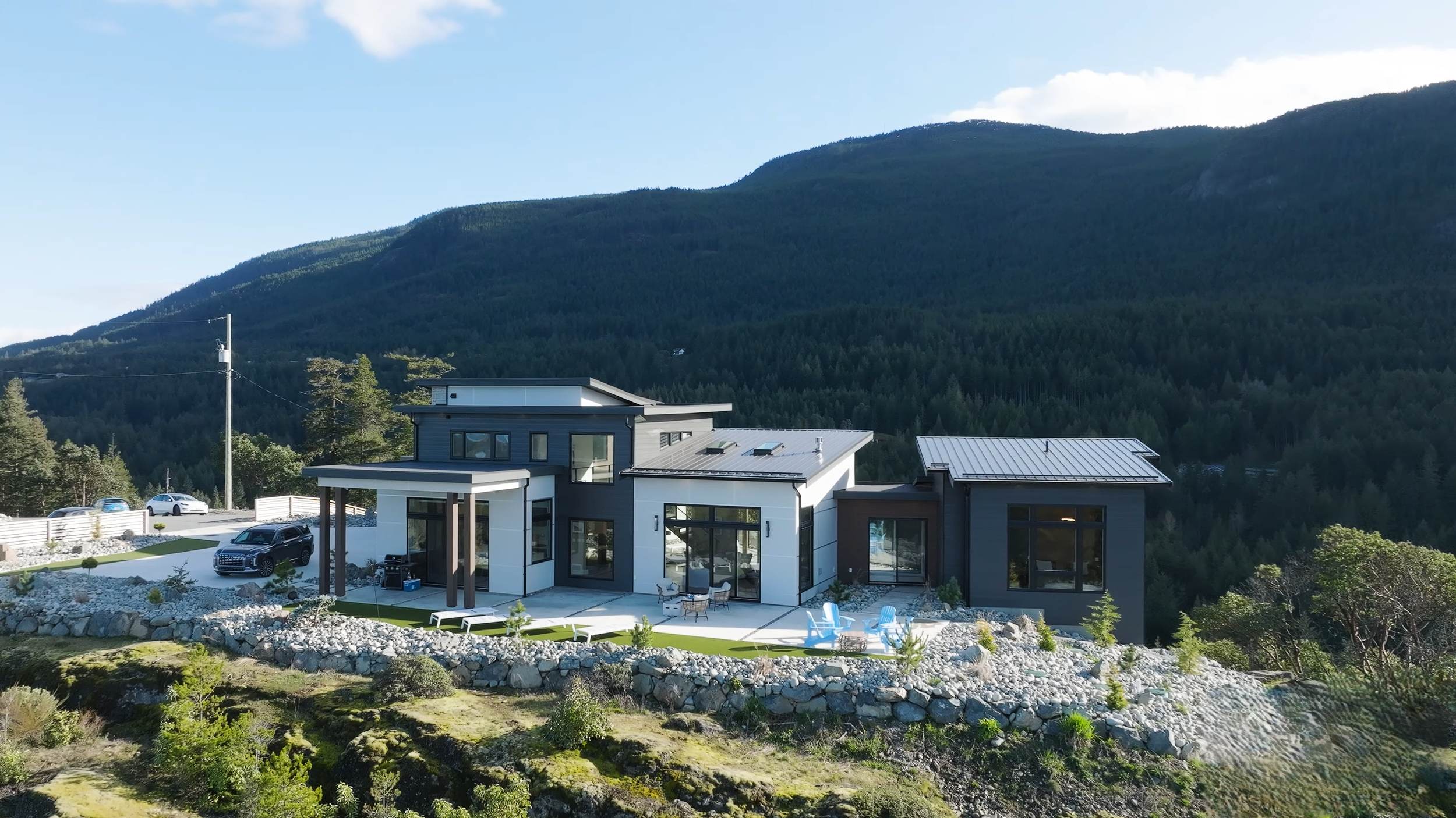 Modern house with large windows and a patio, surrounded by a landscaped yard with outdoor furniture, set against a backdrop of green mountains and blue sky.