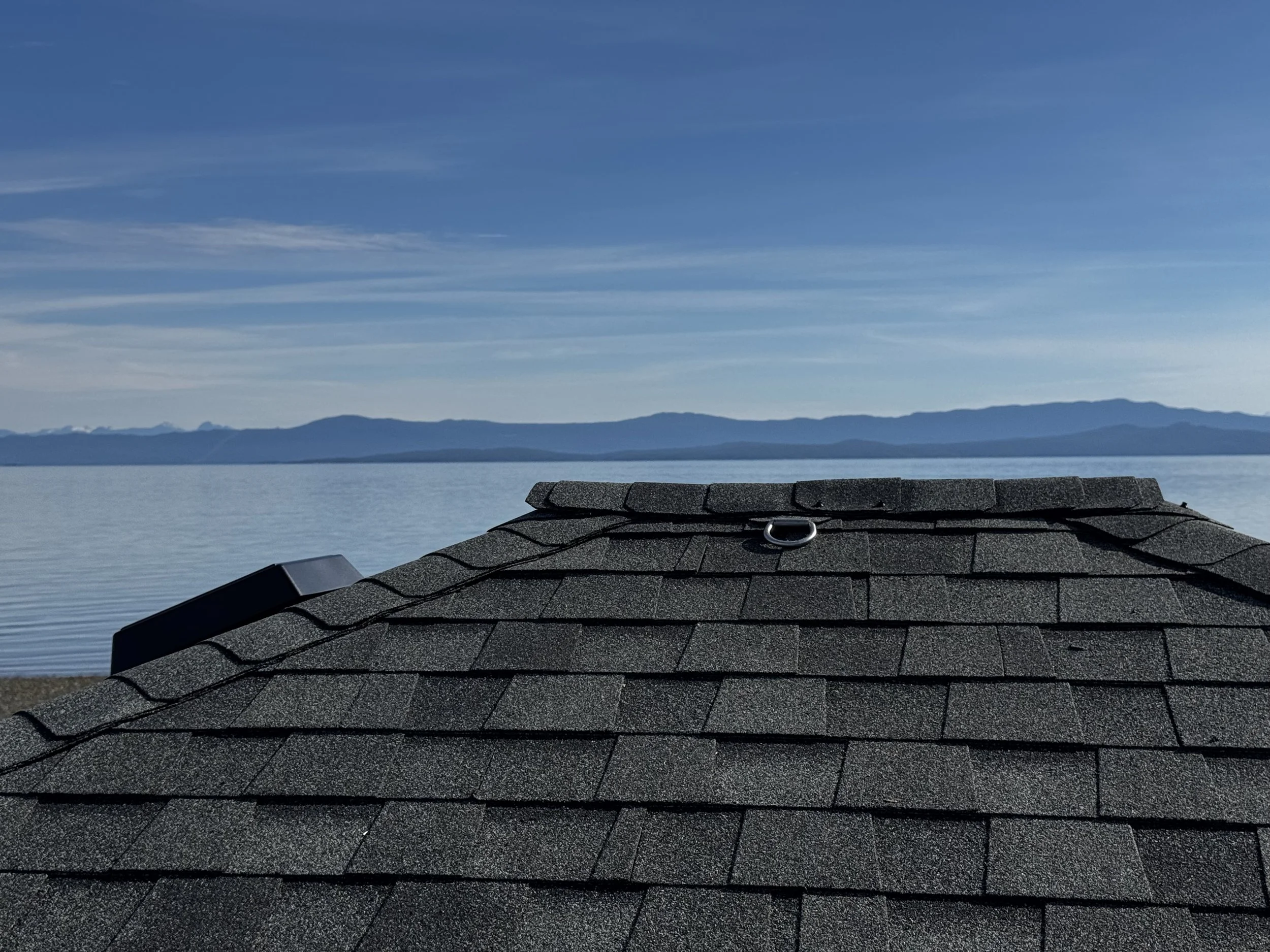 Photo of a rooftop with gray shingles and a metal attachment, overlooking a body of water and distant mountains under a blue sky with wispy clouds.