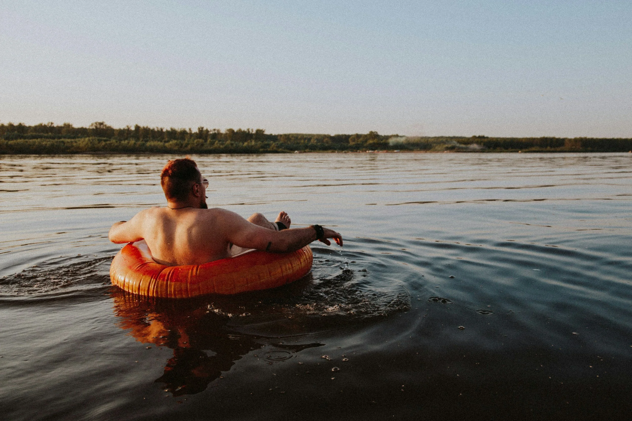 Person lounging on an inflatable tube in calm water with a distant forested shoreline under a clear sky.