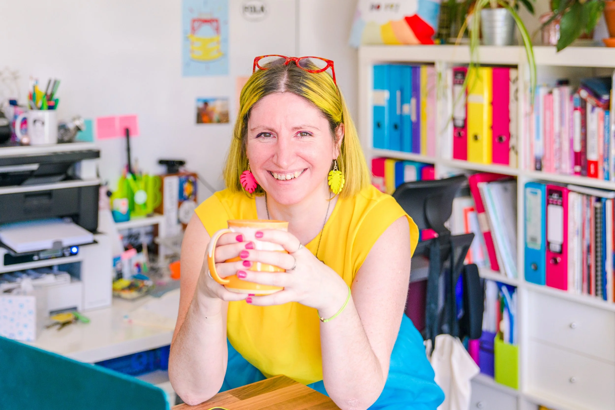 White women with yellow hair, with yellow top, orange glasses on her head, blue skirt. Elbows leaning on a table with in front of a laptop holding a yellow mug. Background is shelves with colourful folders.
