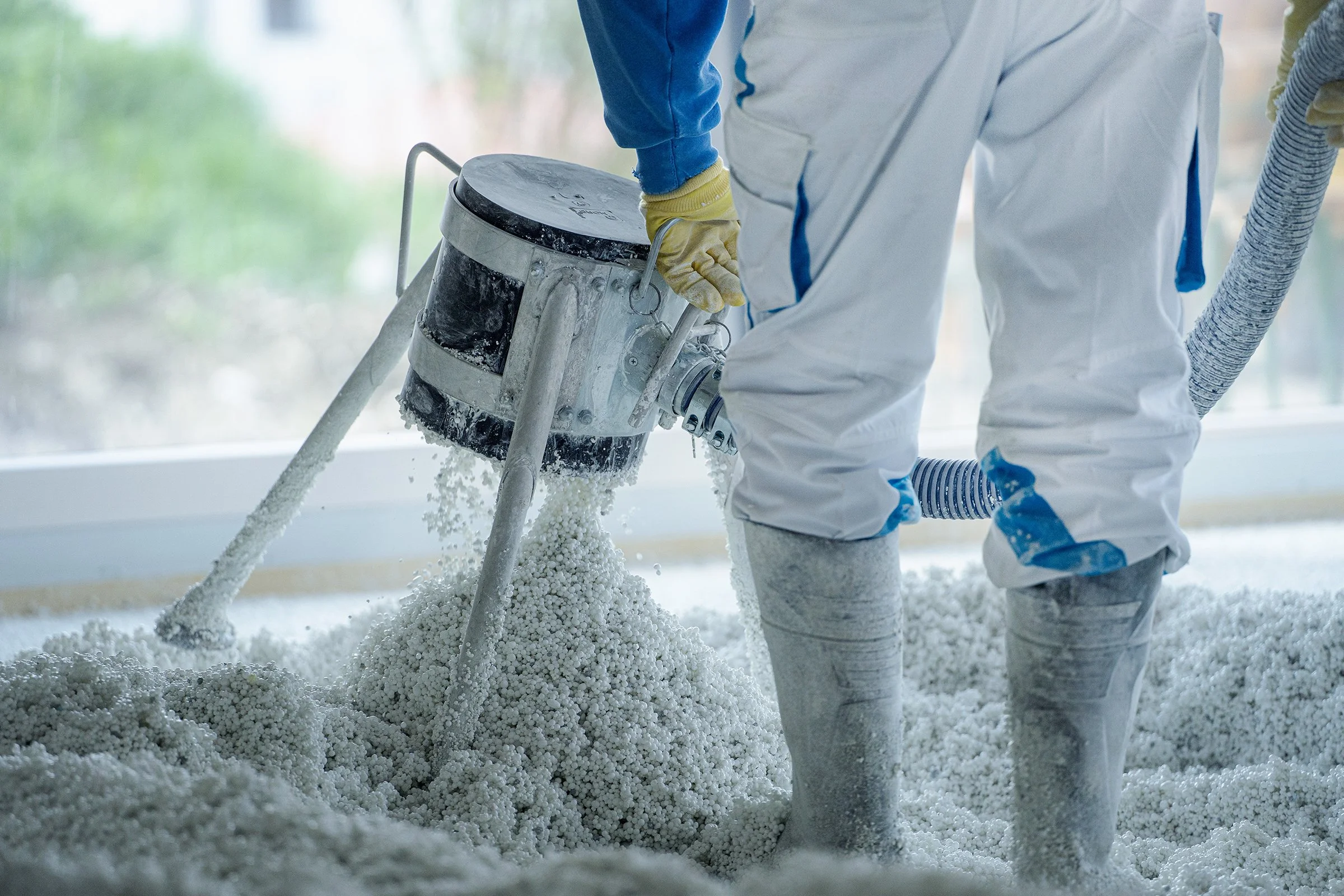 Persoon in witte beschermkleding en gele handschoenen gebruikt mixer om Thermowhite vloerisolatie aan te brengen. Achtergrond is vaag en groen buiten.