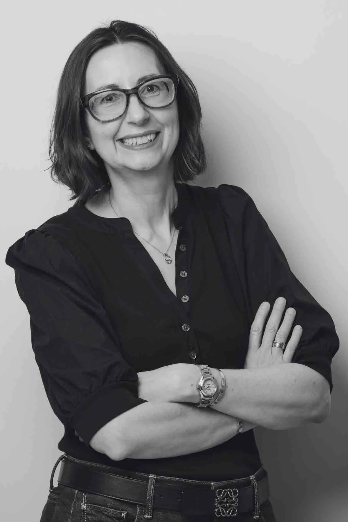 Black and white photo of a woman with shoulder-length hair, wearing glasses, smiling, with arms crossed, dressed in a dark button-up shirt and jeans, standing against a plain background.