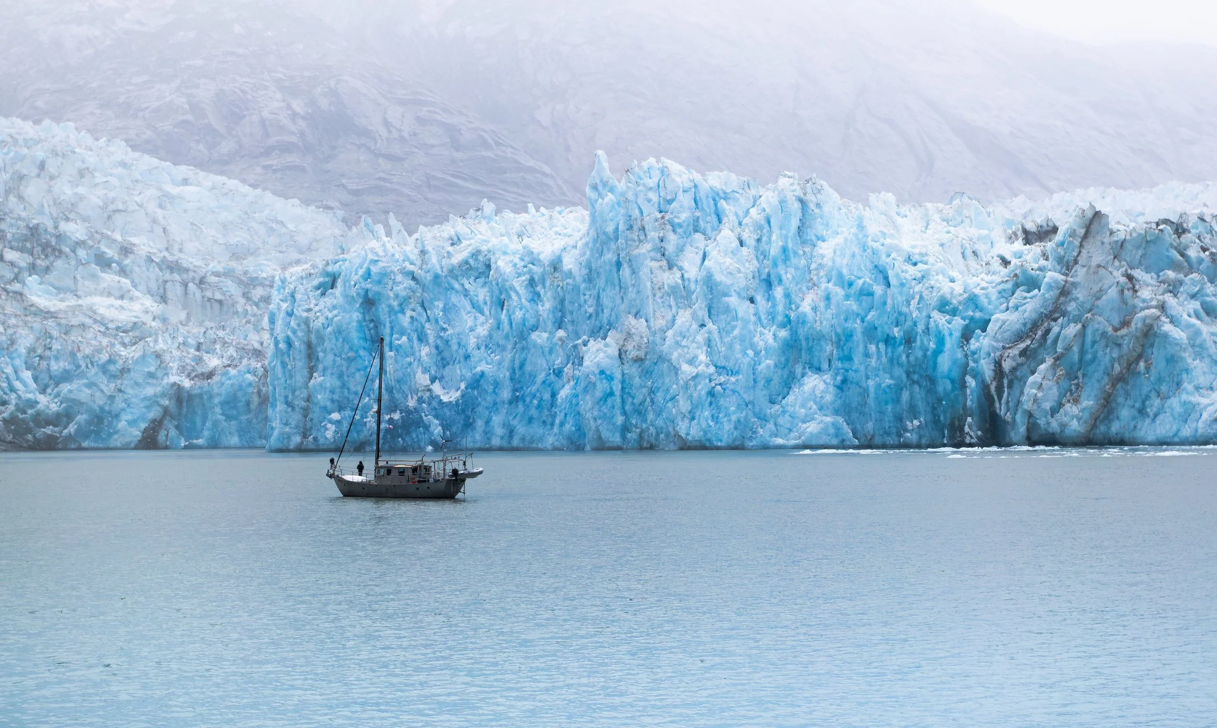 A boat sailing on calm water in front of a large blue glacier with ice formations and mountains in the background.