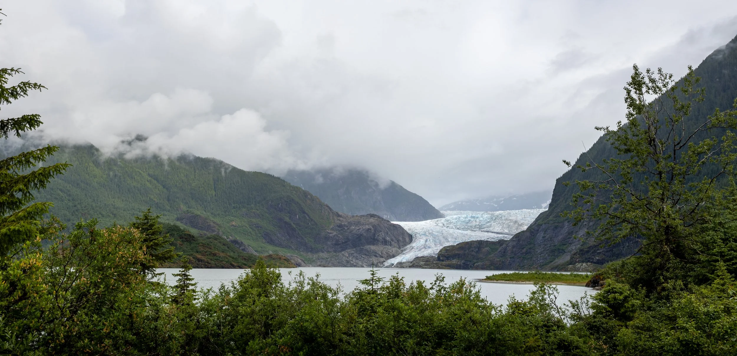 A scenic view of a glacier in a mountainous area with lush green trees in the foreground and low clouds covering the mountain peaks.