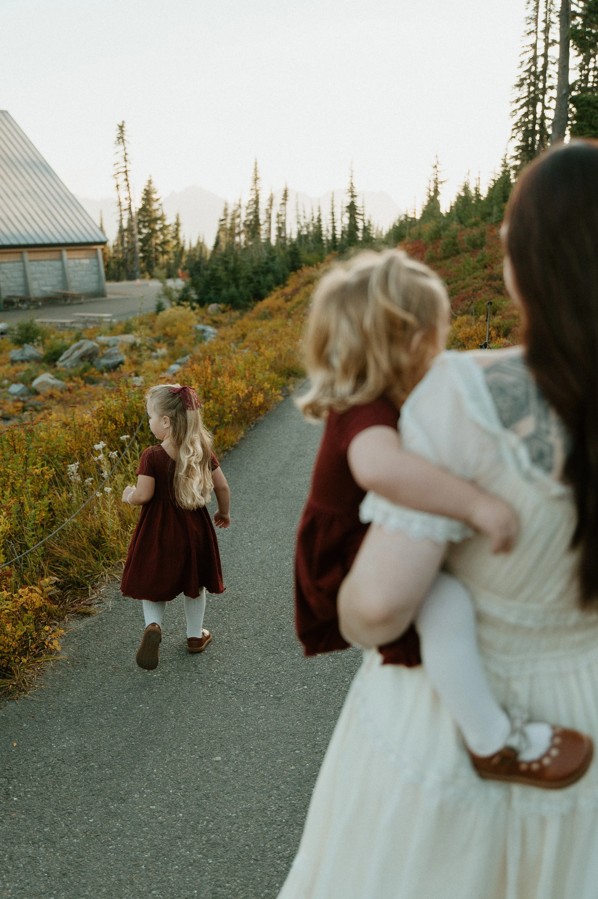 Candid photo of a mother carrying her daughter while her older child explores during a fall family session at Mount Rainier