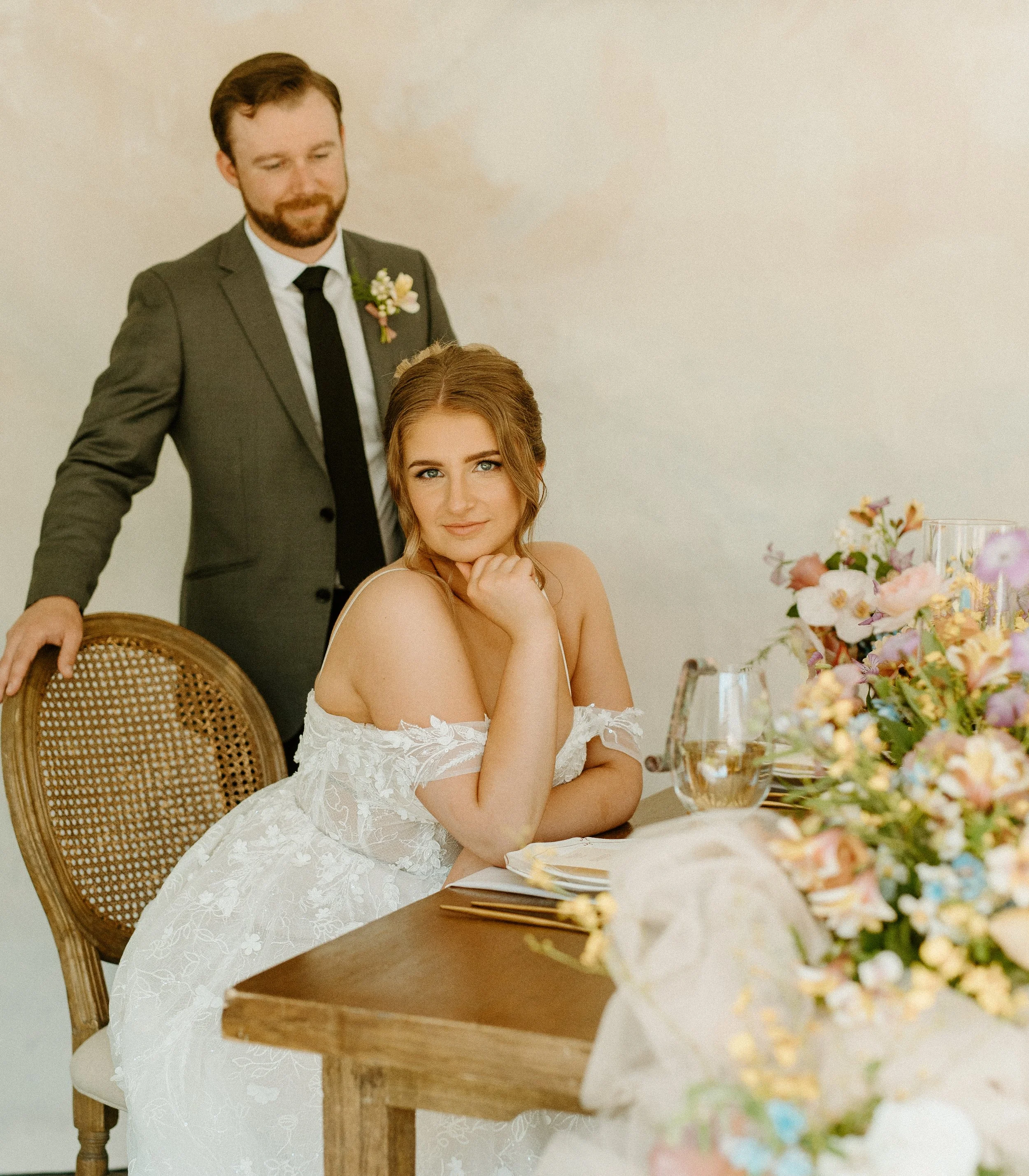 A woman in a white wedding dress sitting at a wooden table with floral arrangements, with a man in a gray suit standing behind her, in an indoor setting.