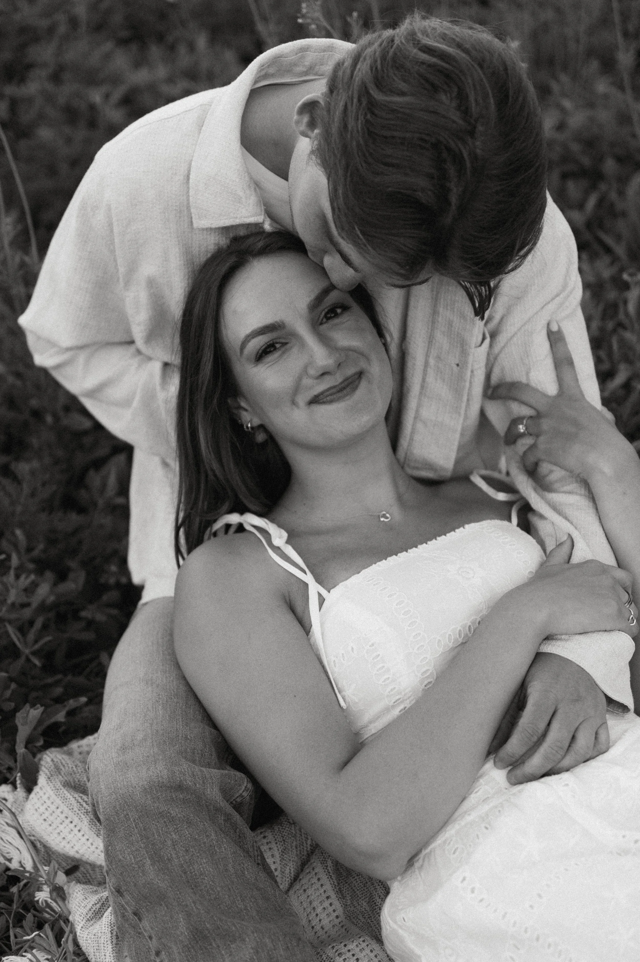 Black-and-white candid of a fiancé kissing his partner’s forehead as they sit together in the wildflowers.