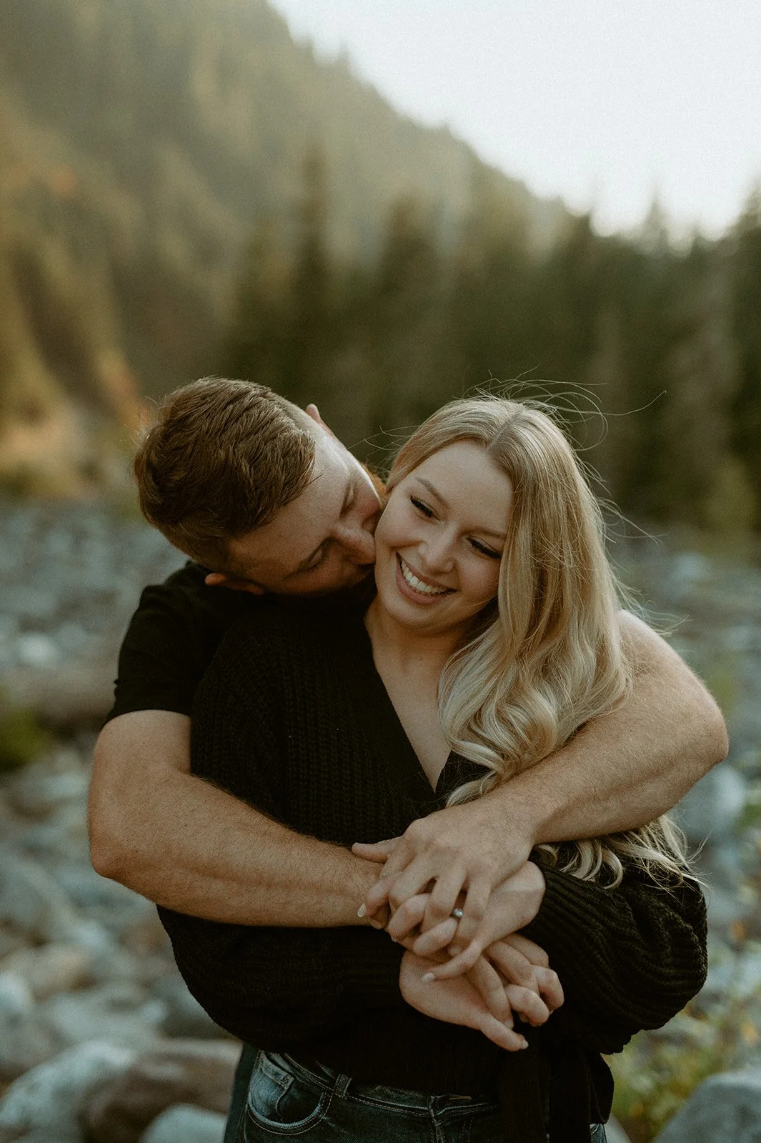 Groom hugging bride from behind near a rocky riverbed during a cozy fall engagement session