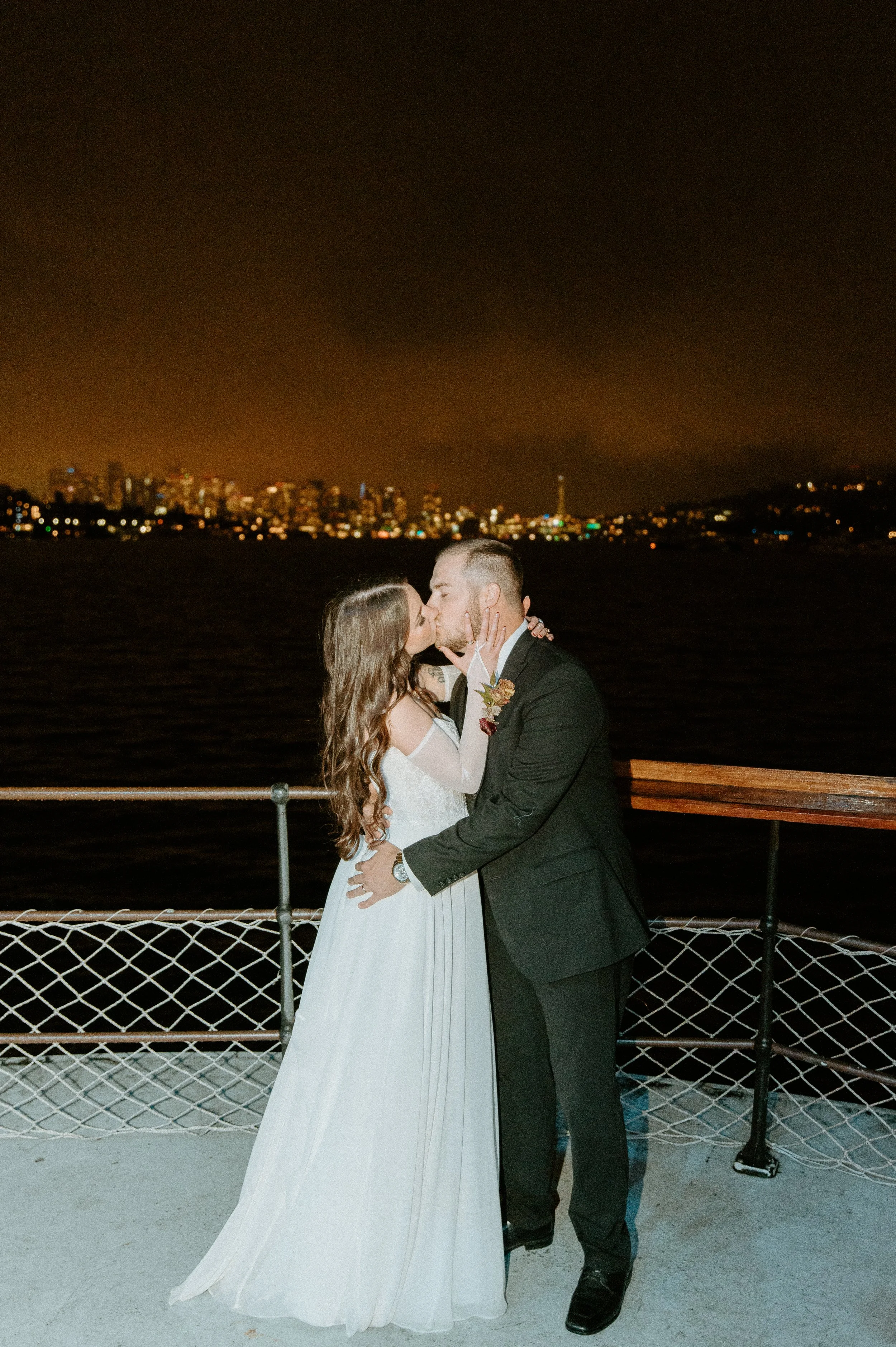 Bride and groom kissing on the deck of a Seattle boat wedding with the city skyline glowing in the background at night