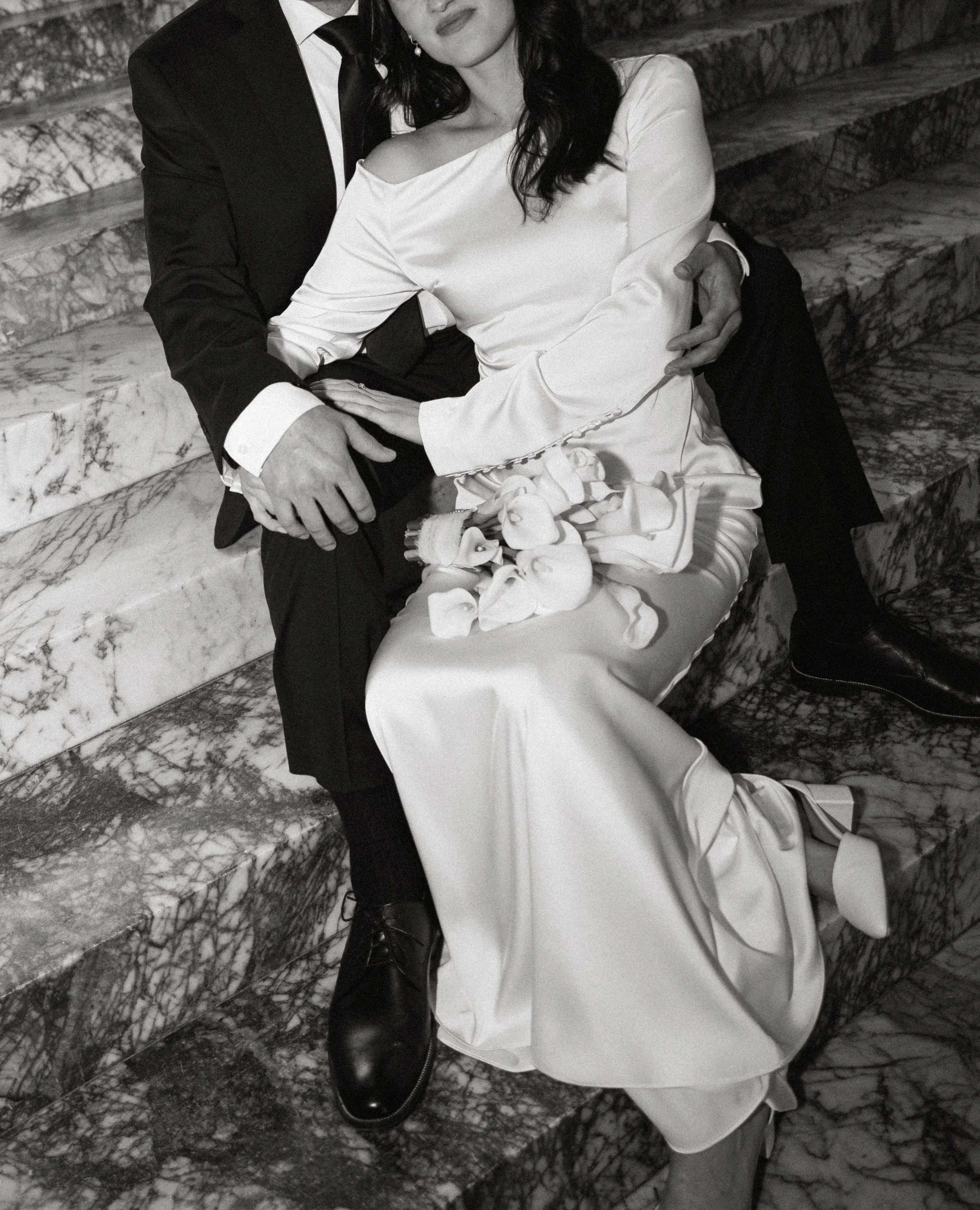 A black-and-white photo of a couple sitting on marble stairs, dressed in formal attire, with the woman holding a bouquet of flowers.