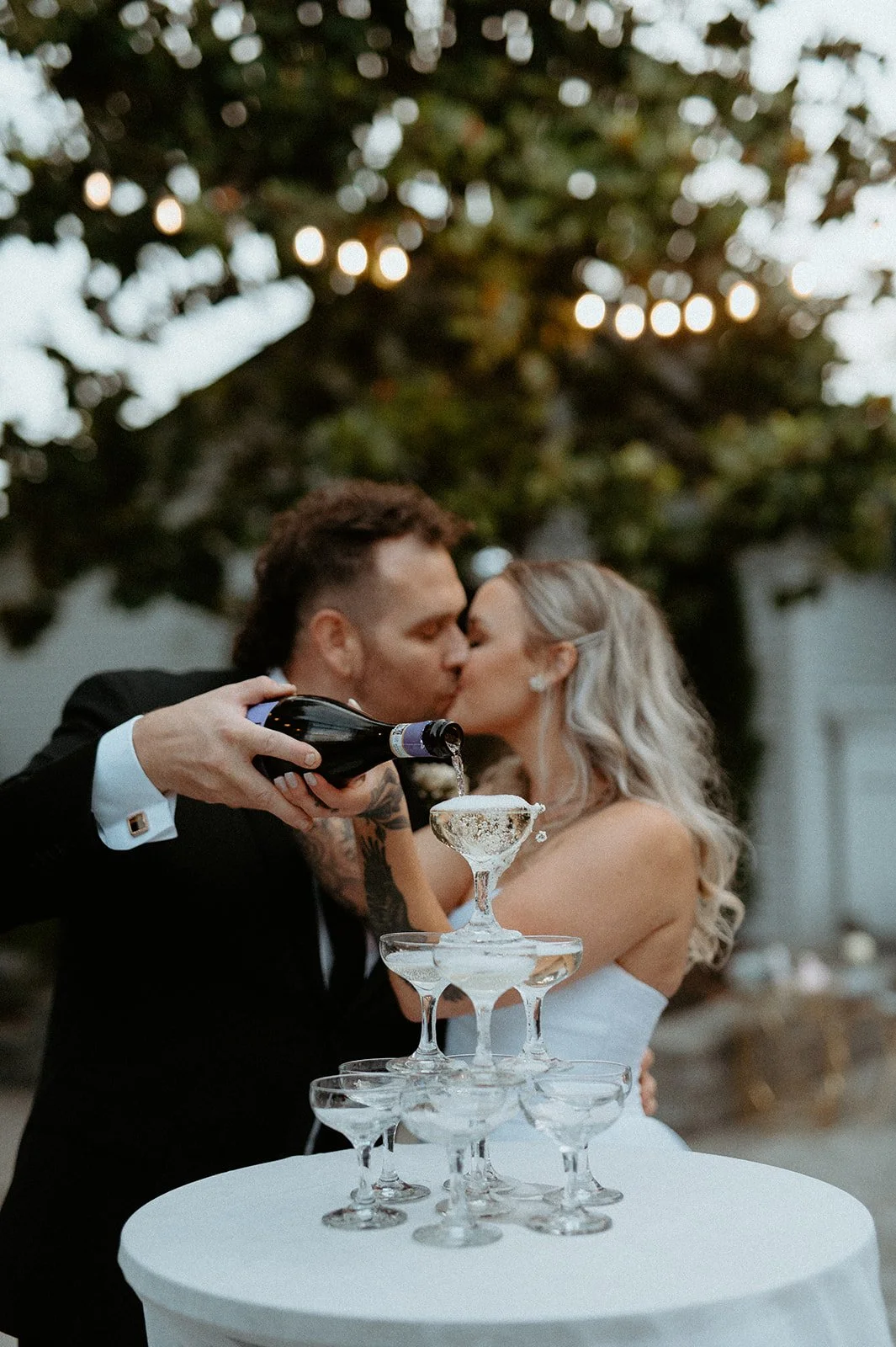 bride + and + groom + kissing + while pouring champagne + over + champagne tower + at + The Orting Manor + wedding + in + Orting WA + with + string lights