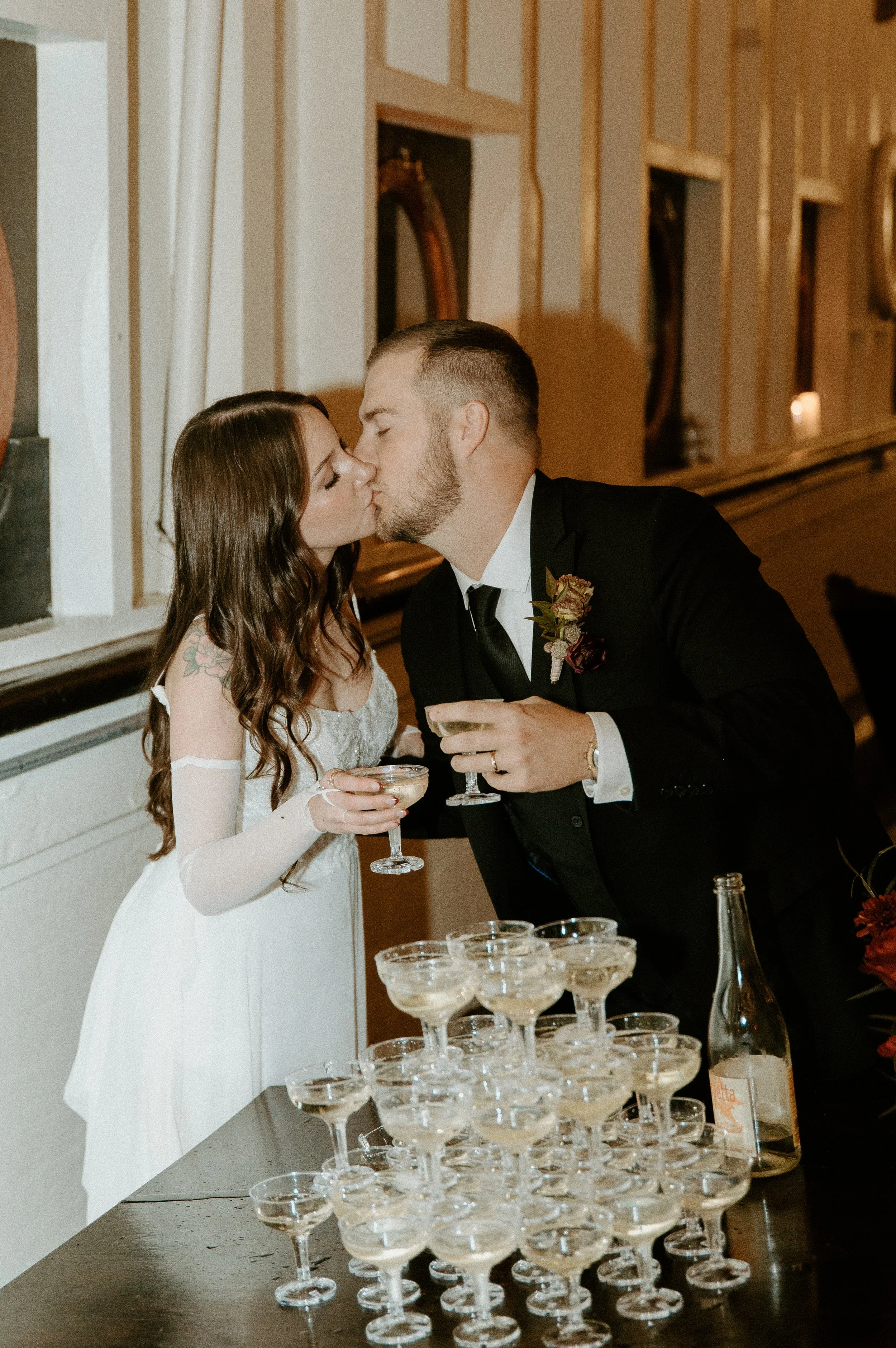 Romantic wedding reception photo of bride and groom kissing while holding champagne coupes beside a champagne tower