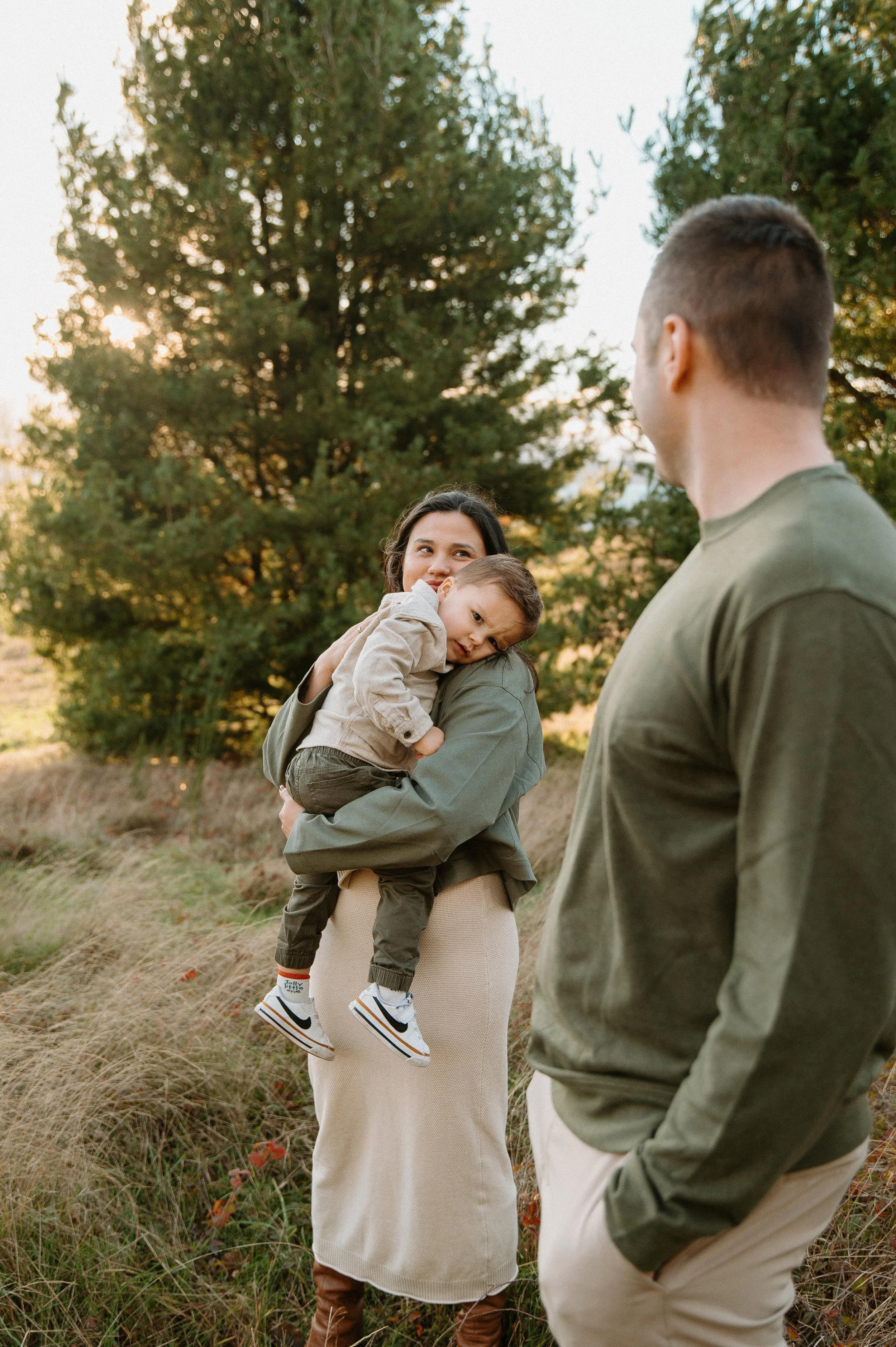 A woman holding a young boy in a grassy outdoor area with trees in the background, and a man standing nearby, all dressed casually.