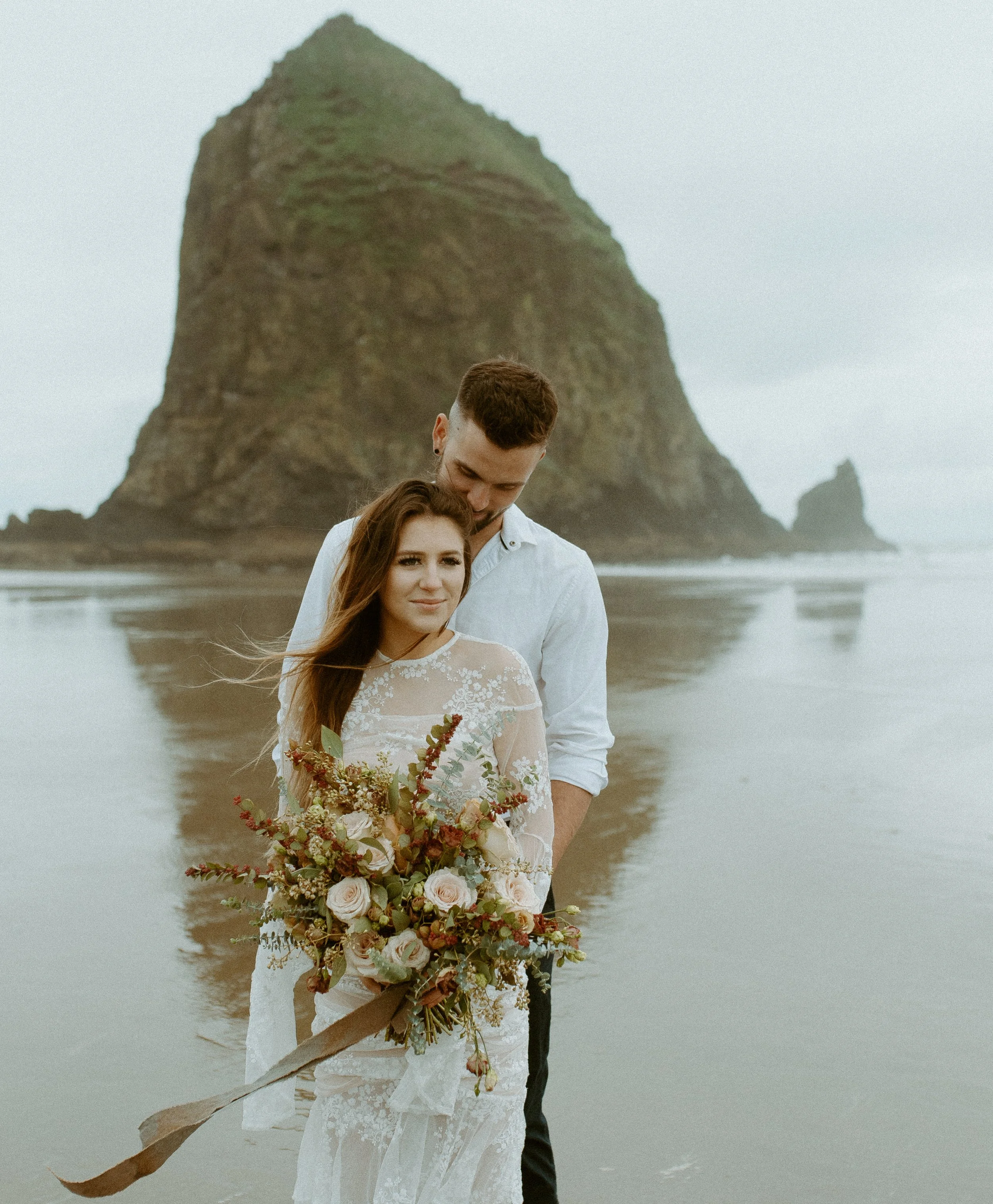 Couple embracing on Cannon Beach Oregon during a moody coastal elopement with Haystack Rock in the background and earthy toned bridal bouquet