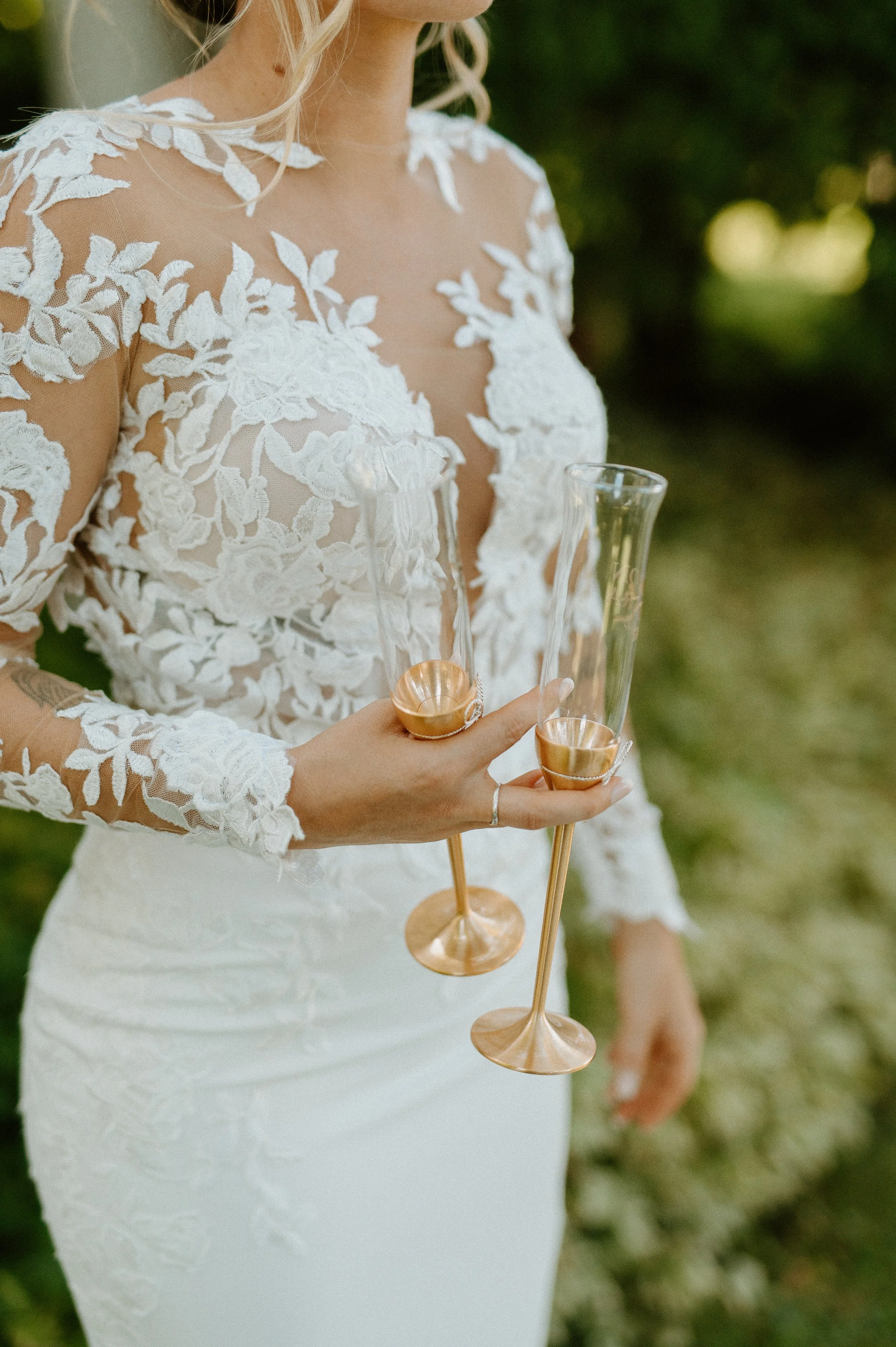 A woman in a white lace wedding dress holding two champagne flutes with gold accents, outdoors with blurred green foliage in the background.