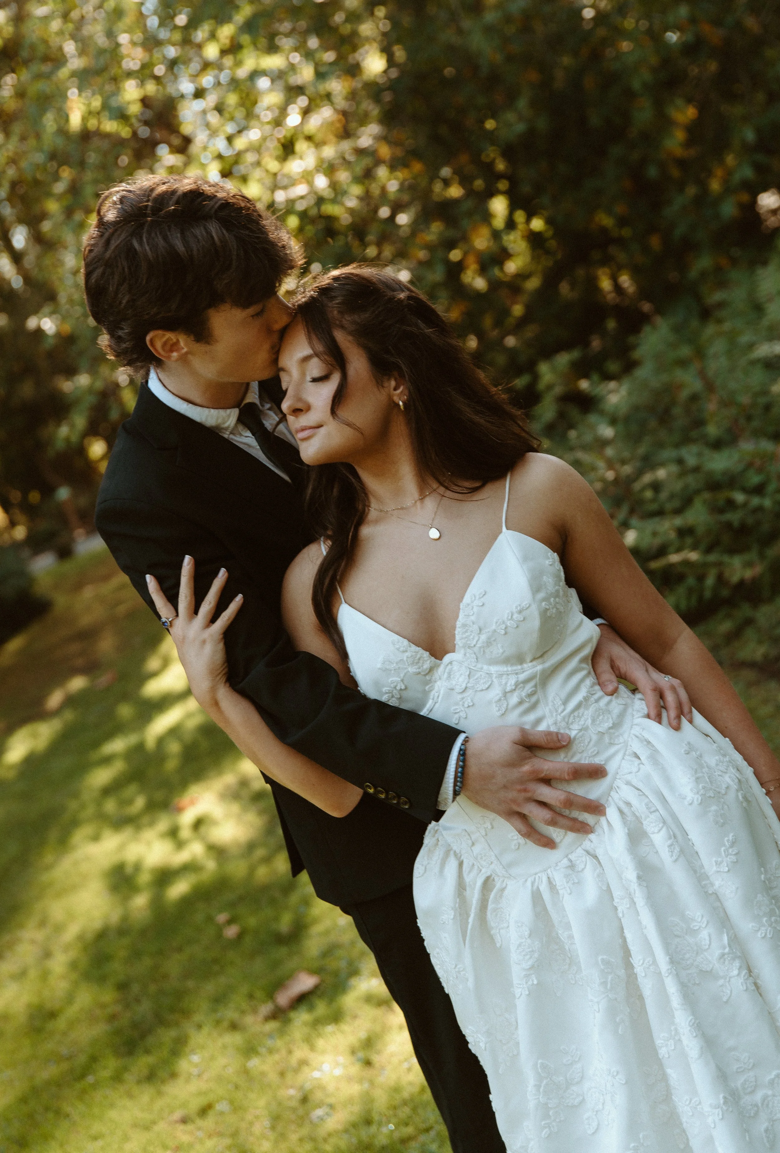 A man in a black suit and a woman in a white wedding dress embrace outdoors, with the man kissing the woman's forehead and her eyes closed, surrounded by trees with sunlight filtering through.