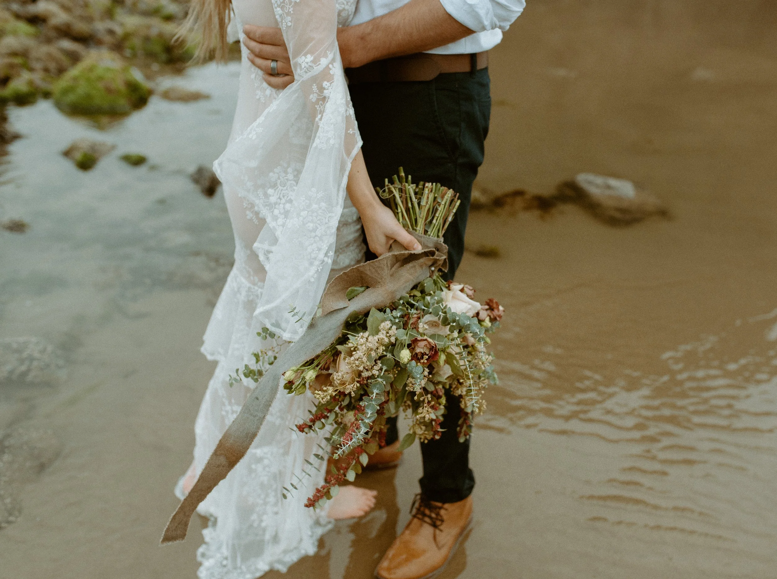 Bride holding earthy toned bridal bouquet while standing barefoot in shallow water on Cannon Beach Oregon