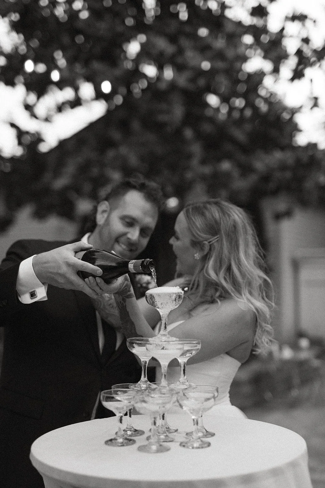 bride + and + groom + pouring champagne + into coupe glass tower + at + The Orting Manor + in + Orting Washington + during + outdoor wedding reception